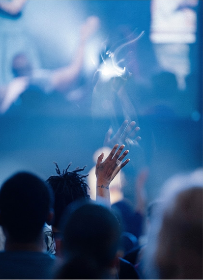 Audience raising hands at a blue-lit concert, with a bright stage light in the background