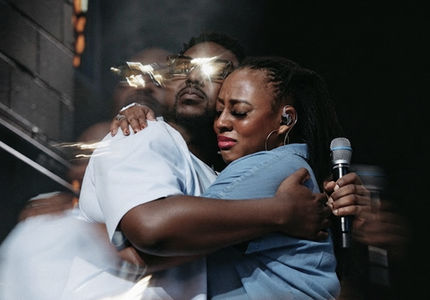 Two people hugging on stage, one holding a microphone, with bright lights in the background