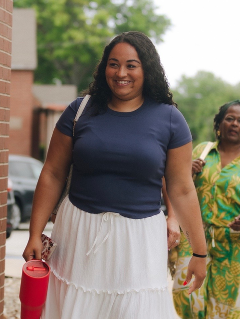 Woman walking outdoors in a blue top and white skirt, carrying a red water bottle