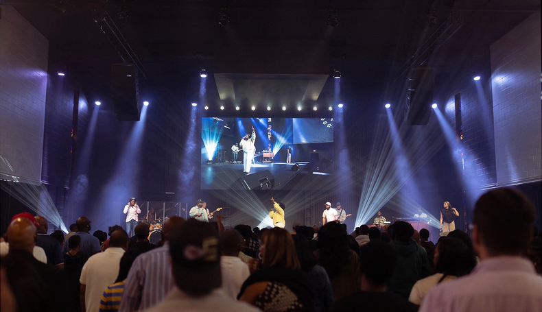 Concert crowd facing a brightly lit stage with a large screen and purple spotlights