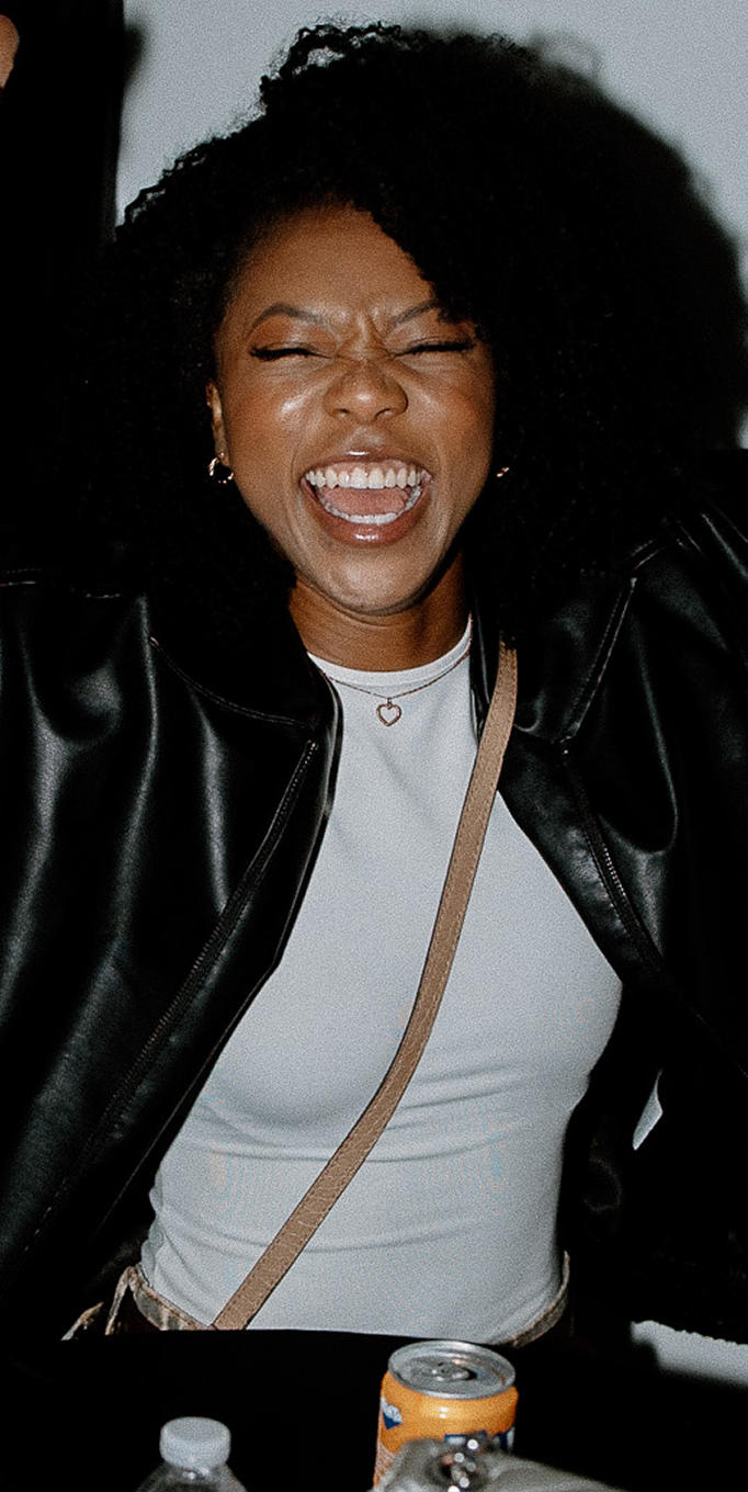 Smiling person in a black jacket and white top at a table with a drink can