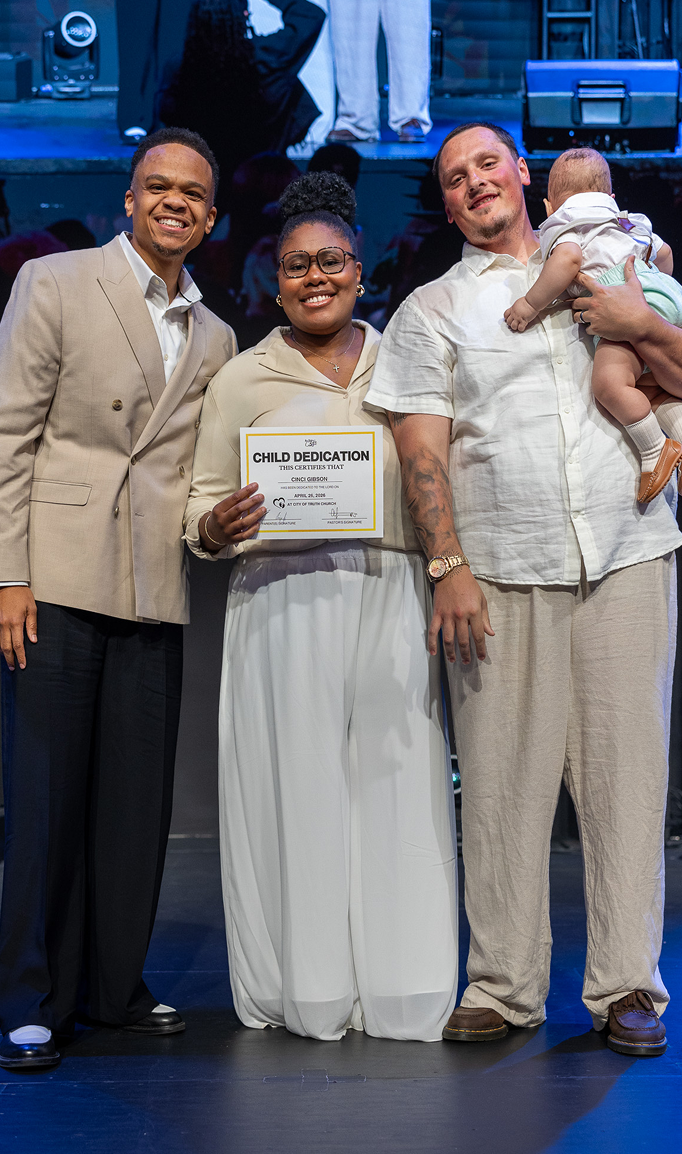 Three people posing onstage; center person holds a certificate, and the man on right holds a baby.