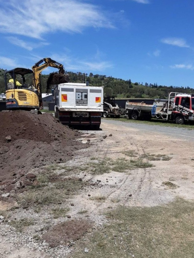 Yellow excavator loading a white BC truck with dark soil on a sunny day. Other trucks parked nearby — BC Earthmoving and Tipper Hire in Gympie, QLD