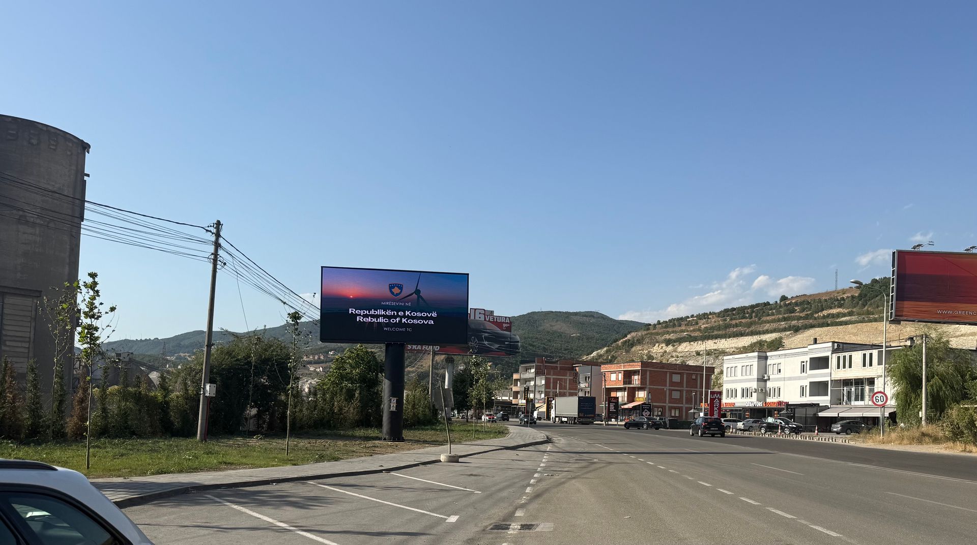 Billboard in a town square displays a sunset with text. Road in foreground, buildings and hills in background.