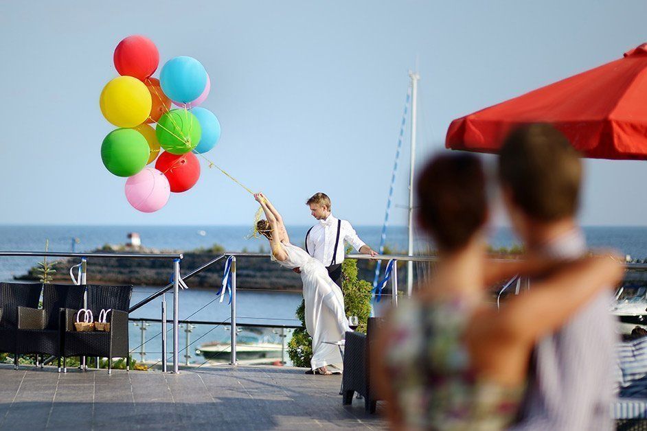 Woman held aloft by colorful balloons, man watching on a waterfront terrace.