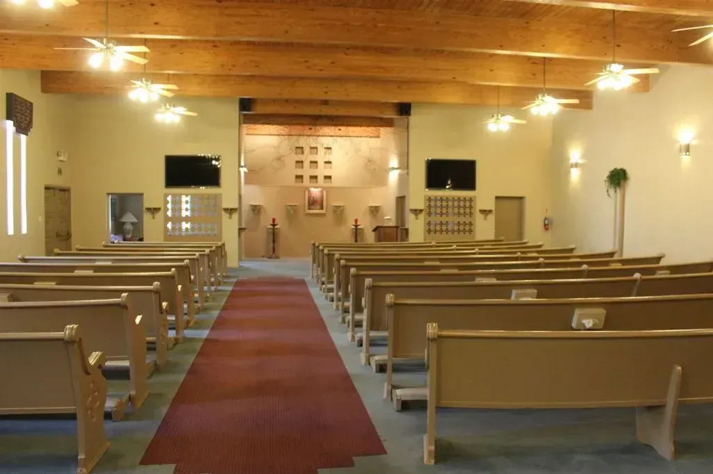 Interior of a chapel with rows of wooden pews, red carpet aisle, and light-colored walls.