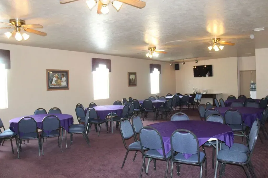 A room with round tables covered in purple tablecloths, blue chairs, ceiling fans, and framed art.