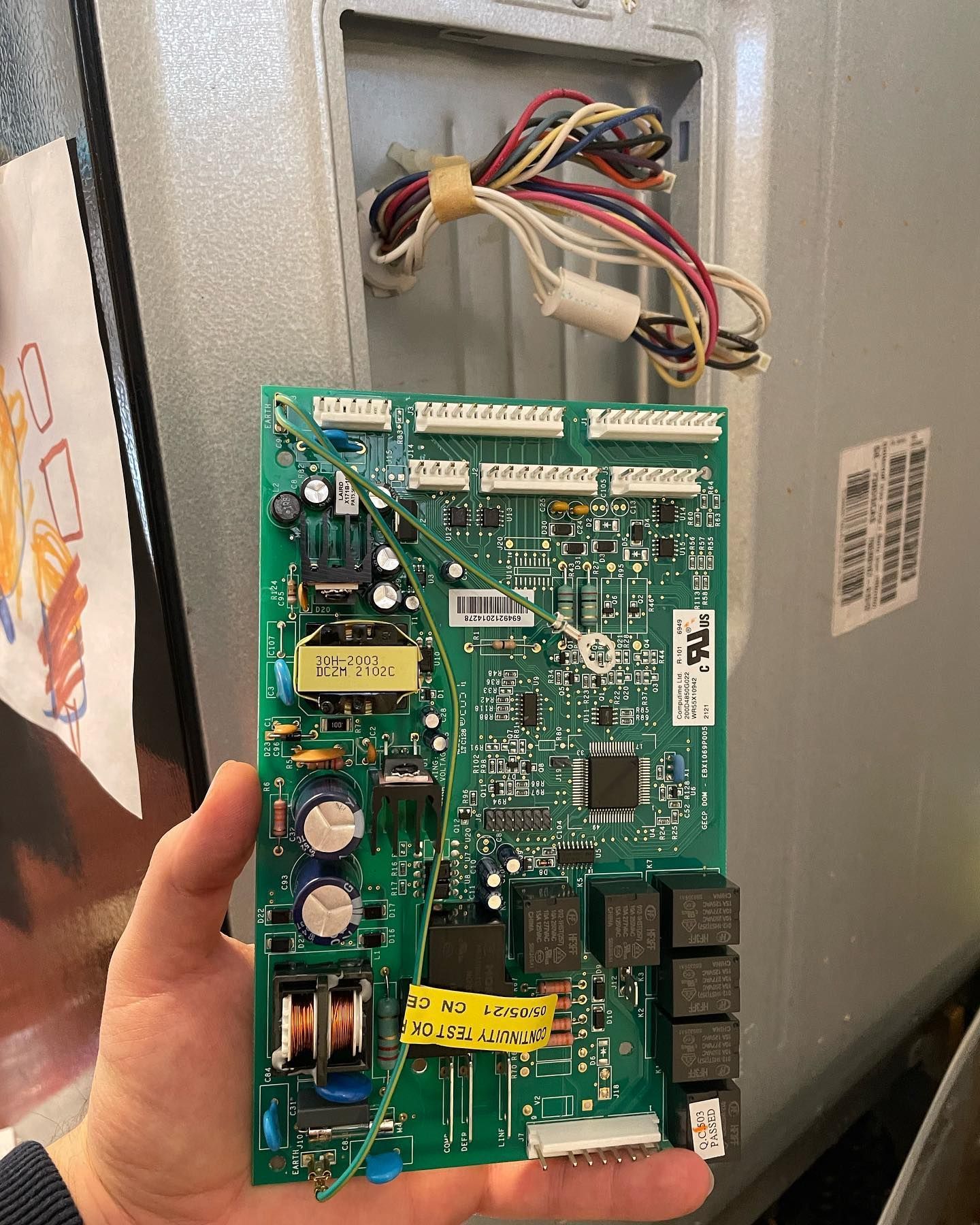 A person is holding a motherboard in their hand in front of a refrigerator.