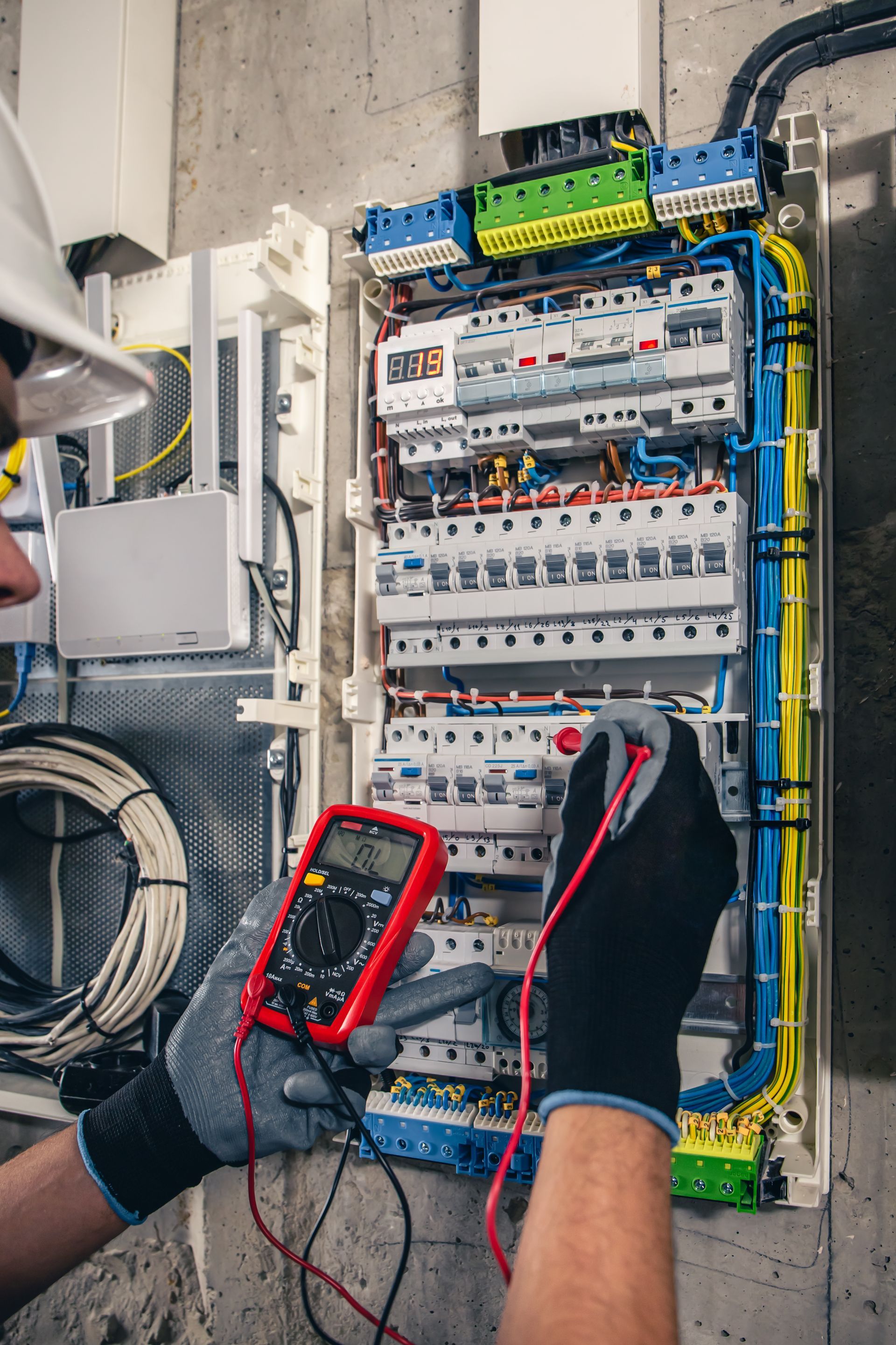 Electrician testing a wall-mounted breaker panel with a multimeter and gloved hands.