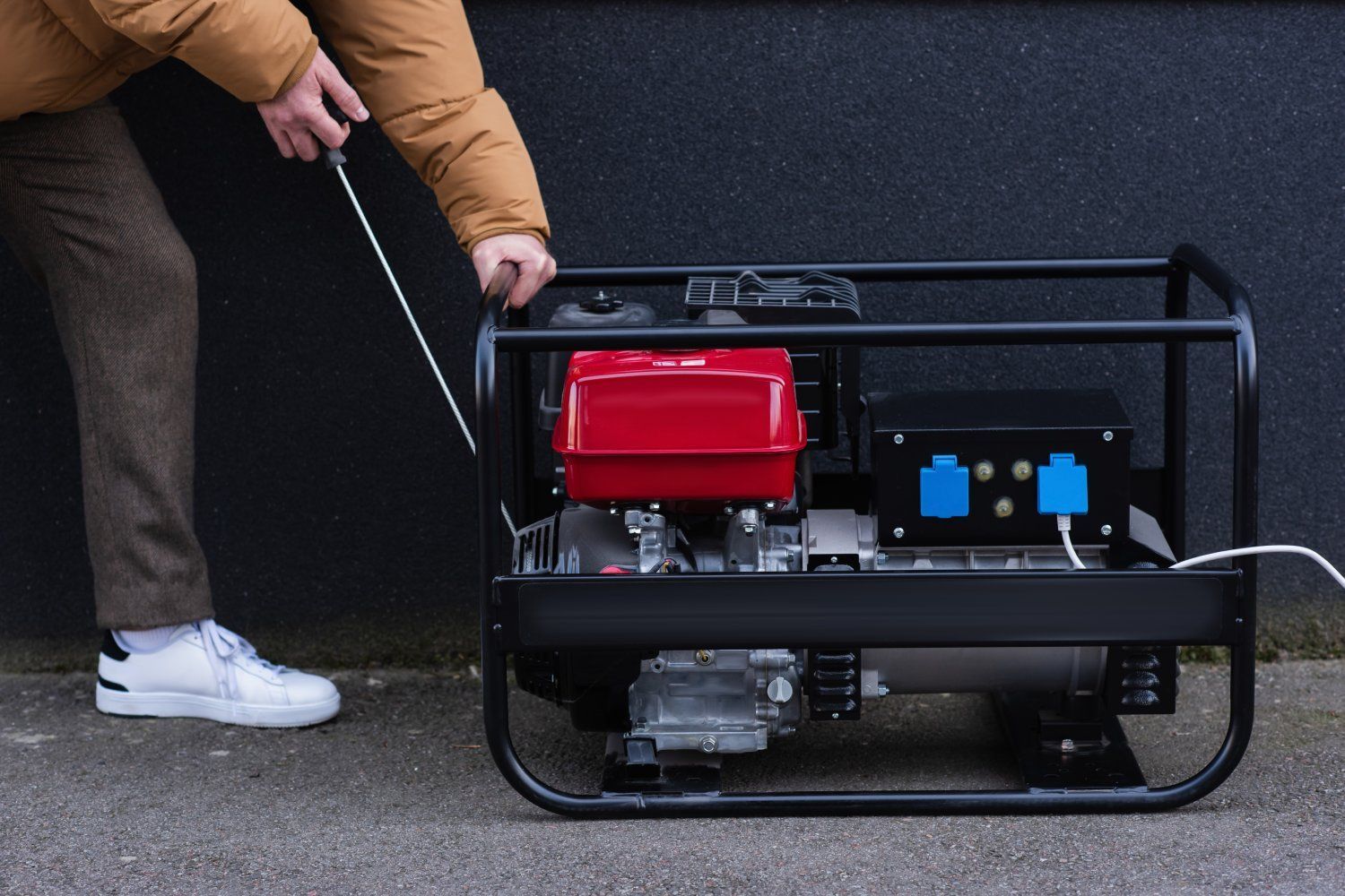 Person starting a red-and-black portable generator with a pull cord outdoors.