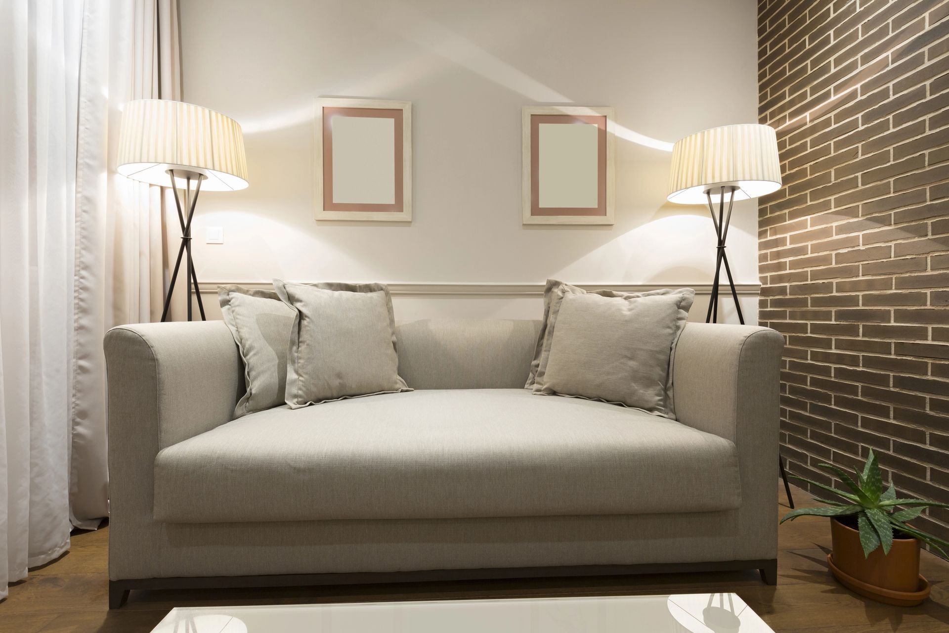 Accogliente soggiorno beige con divano, lampade da terra, quadri e una parete con rivestimento in mattoni a vista.