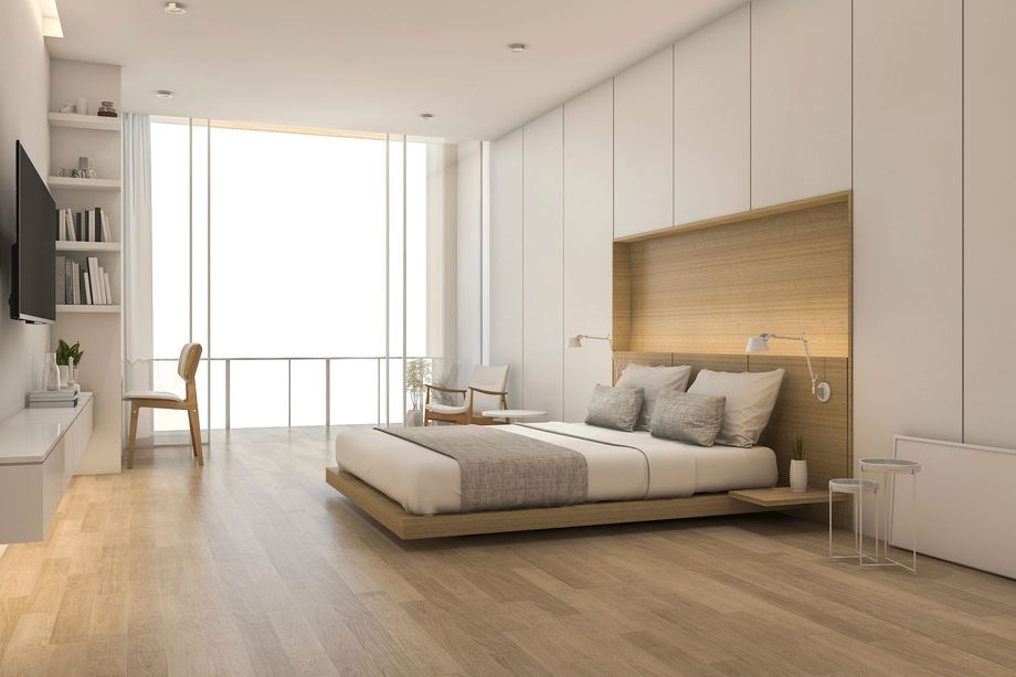 Camera da letto minimalista e luminosa con letto basso, pavimento in legno, pareti bianche e scaffalatura a muro accanto a una grande finestra.
