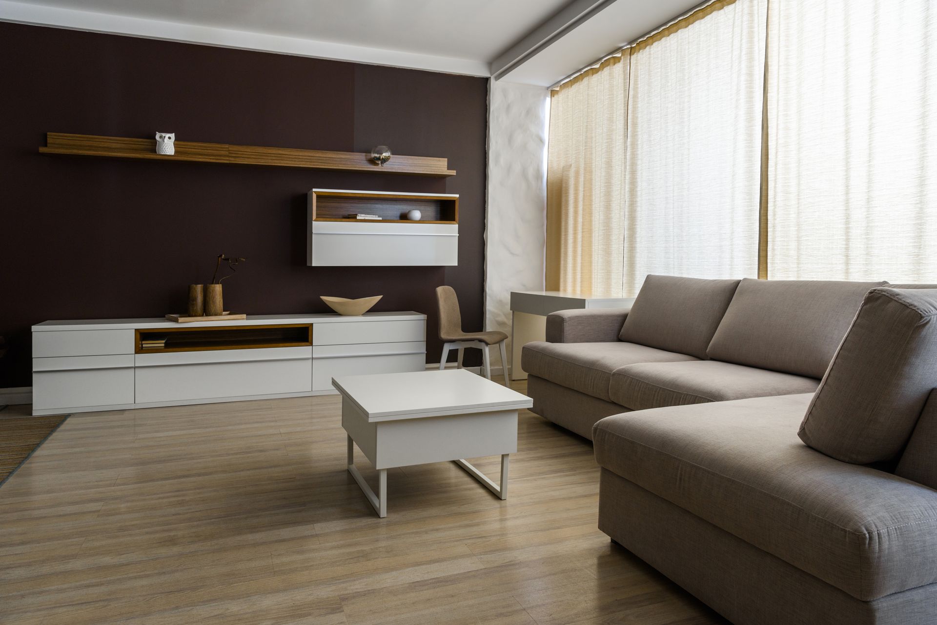 Soggiorno moderno con divano componibile grigio, tavolino da caffè bianco e mobile TV accanto a grandi finestre con tende.
