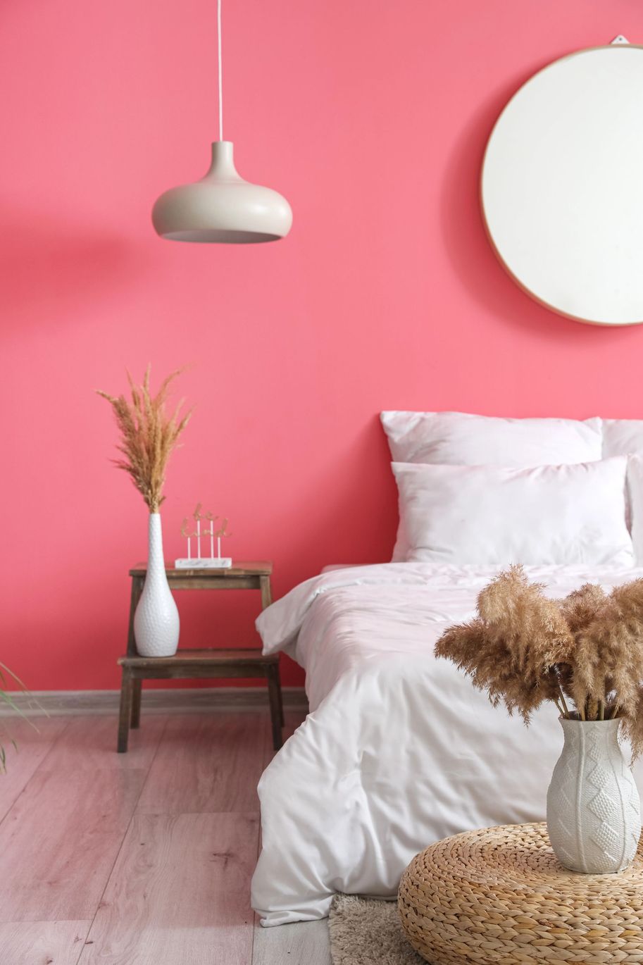 Camera da letto rosa con letto bianco, lampada a sospensione, specchio rotondo e decorazioni intrecciate sul pavimento.