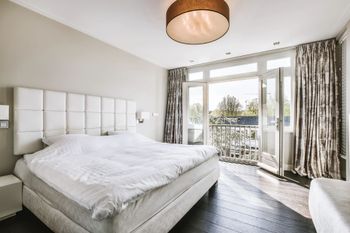 Camera da letto luminosa con letto bianco, porte finestre che danno sul balcone e tende decorate con vista sugli alberi.