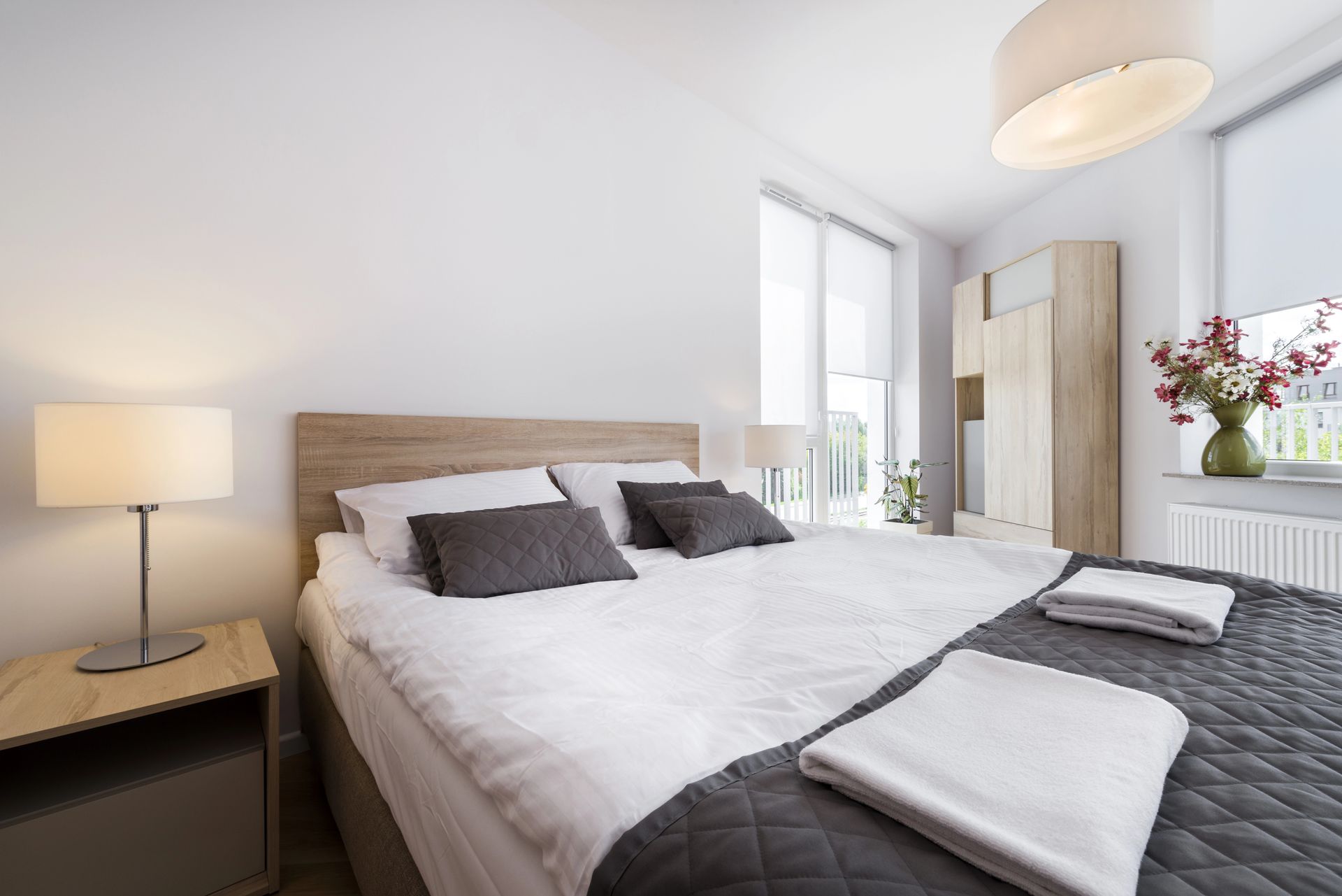 Camera da letto luminosa con letto, lampada da comodino, armadio e finestra con fiori; asciugamani piegati ai piedi del letto.