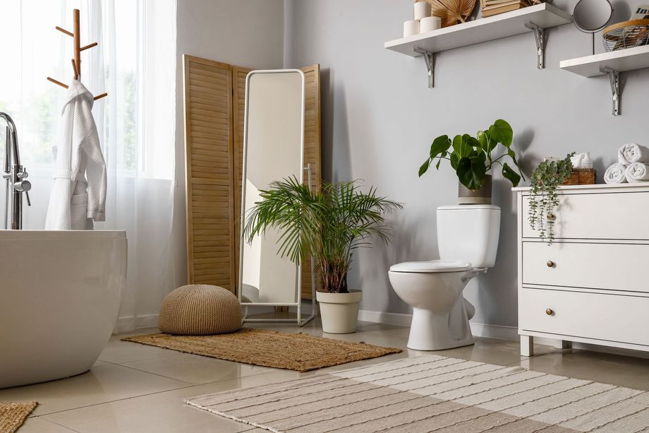 Bagno luminoso e minimalista con vasca bianca, WC, tappeto in vimini, piante e paravento a persiana aperto vicino a una finestra.