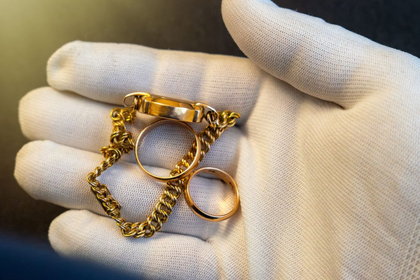 White-gloved hand holding two gold chains and a gold ring on a light background