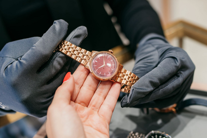 Gold wristwatch with pink face being held by gloved hands over a table