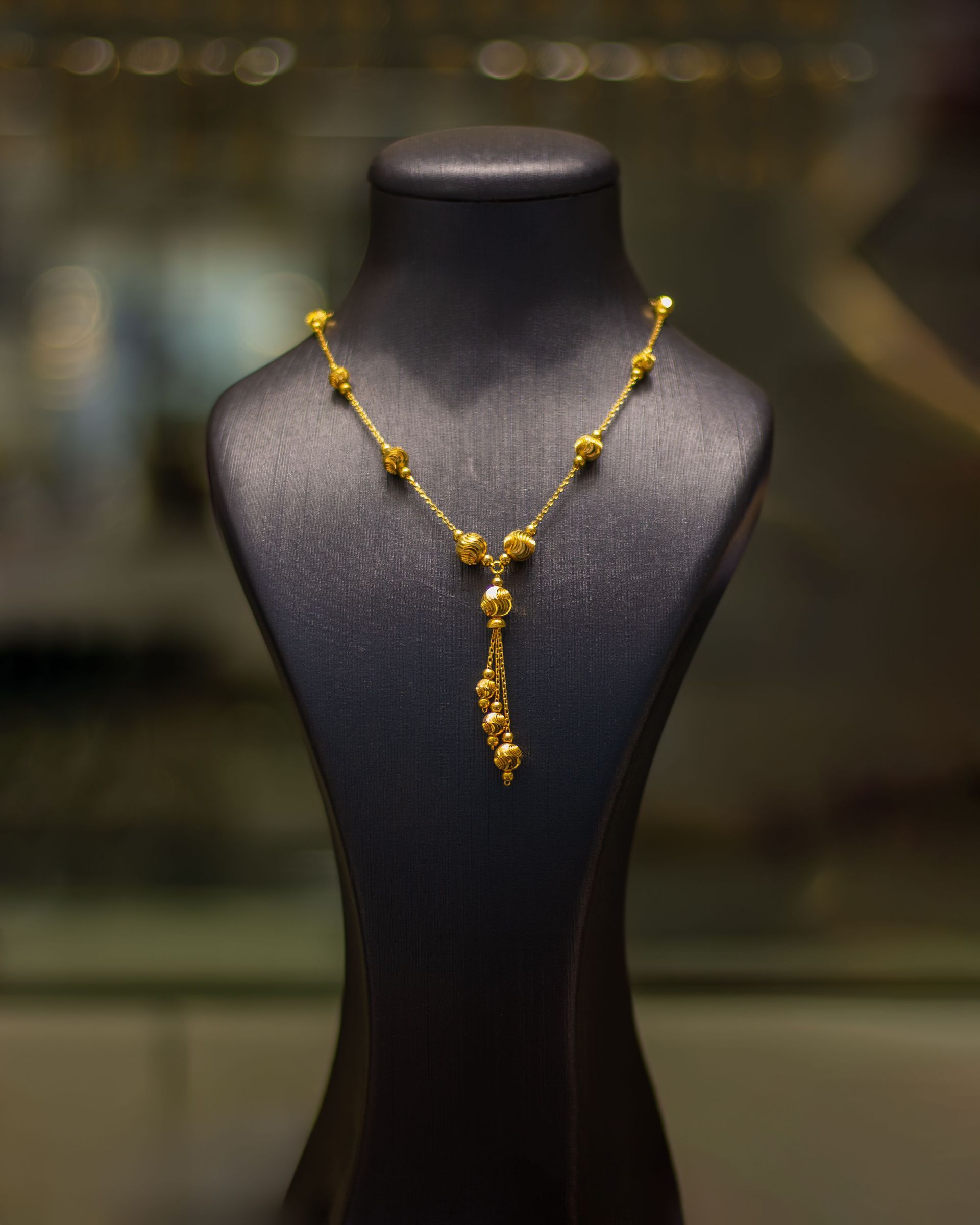 Gold beaded necklace on a black display bust, centered against a blurred background.
