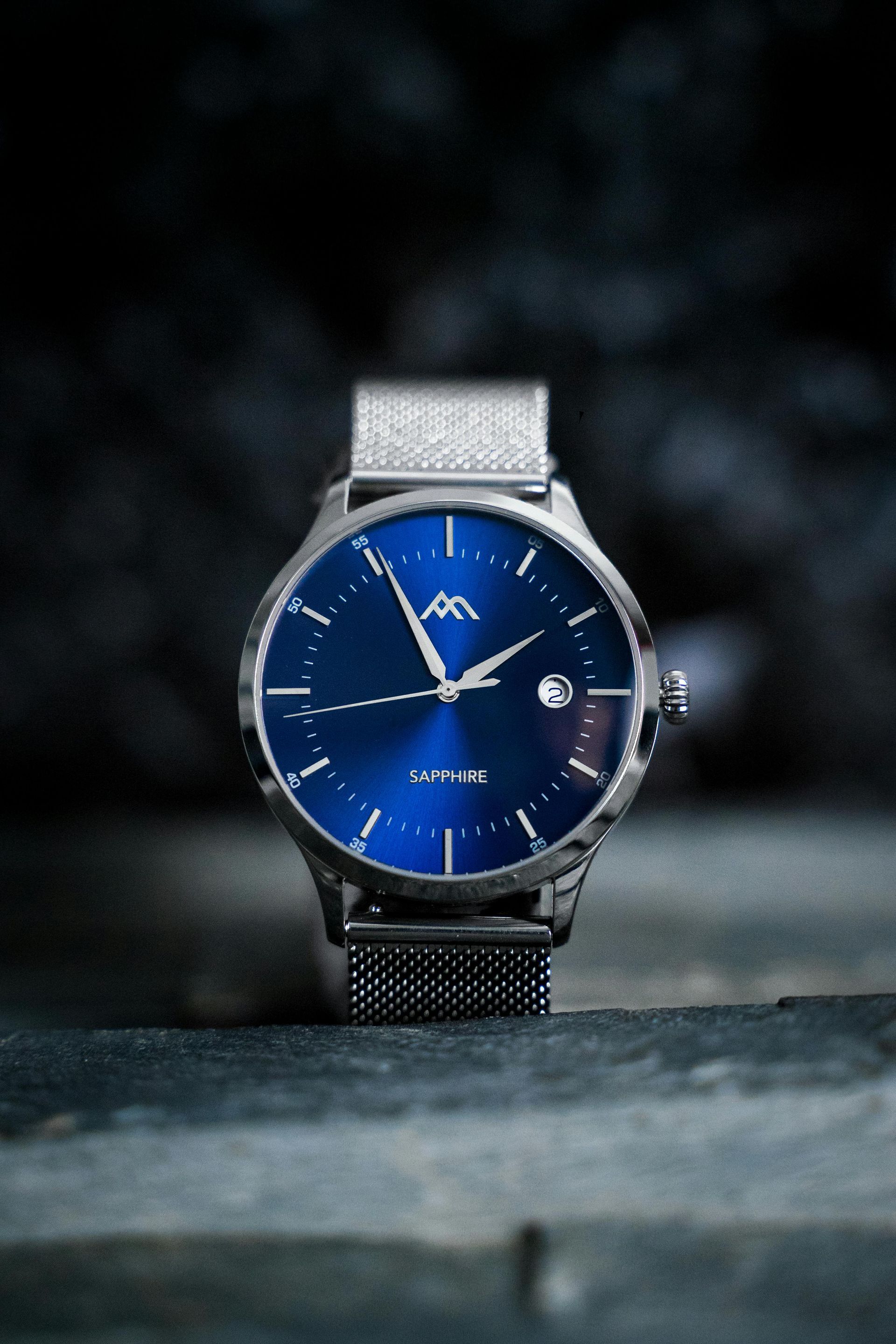 Blue-faced wristwatch with silver mesh band on a dark surface, set against a blurred background
