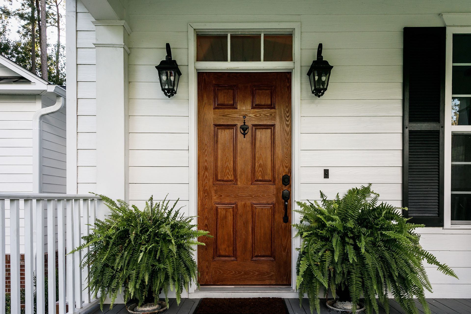 Wooden front door with ferns, black sconces, and white siding.