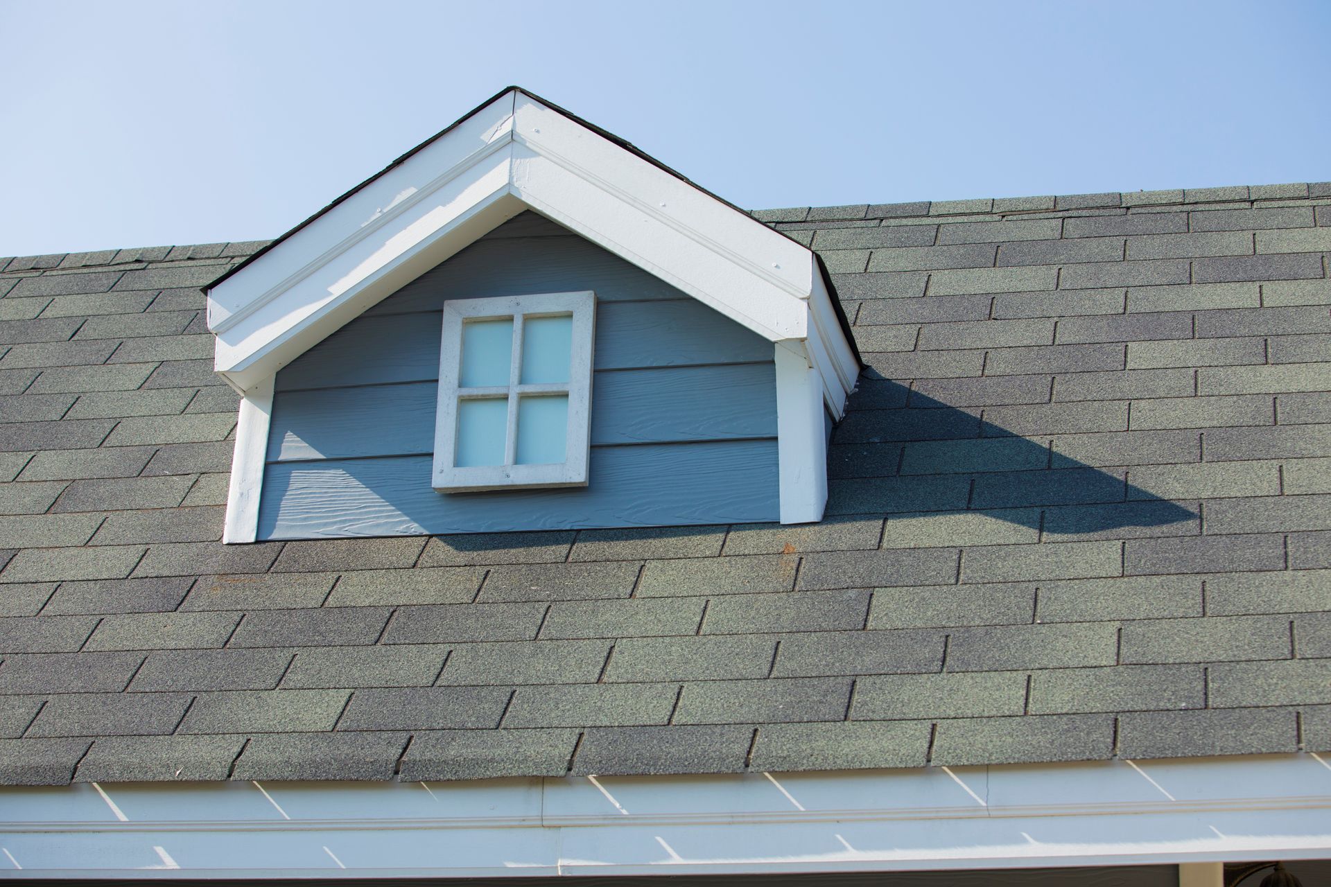 Dormer window on a gray shingled roof with white trim and a blue siding. Bright, sunny day.