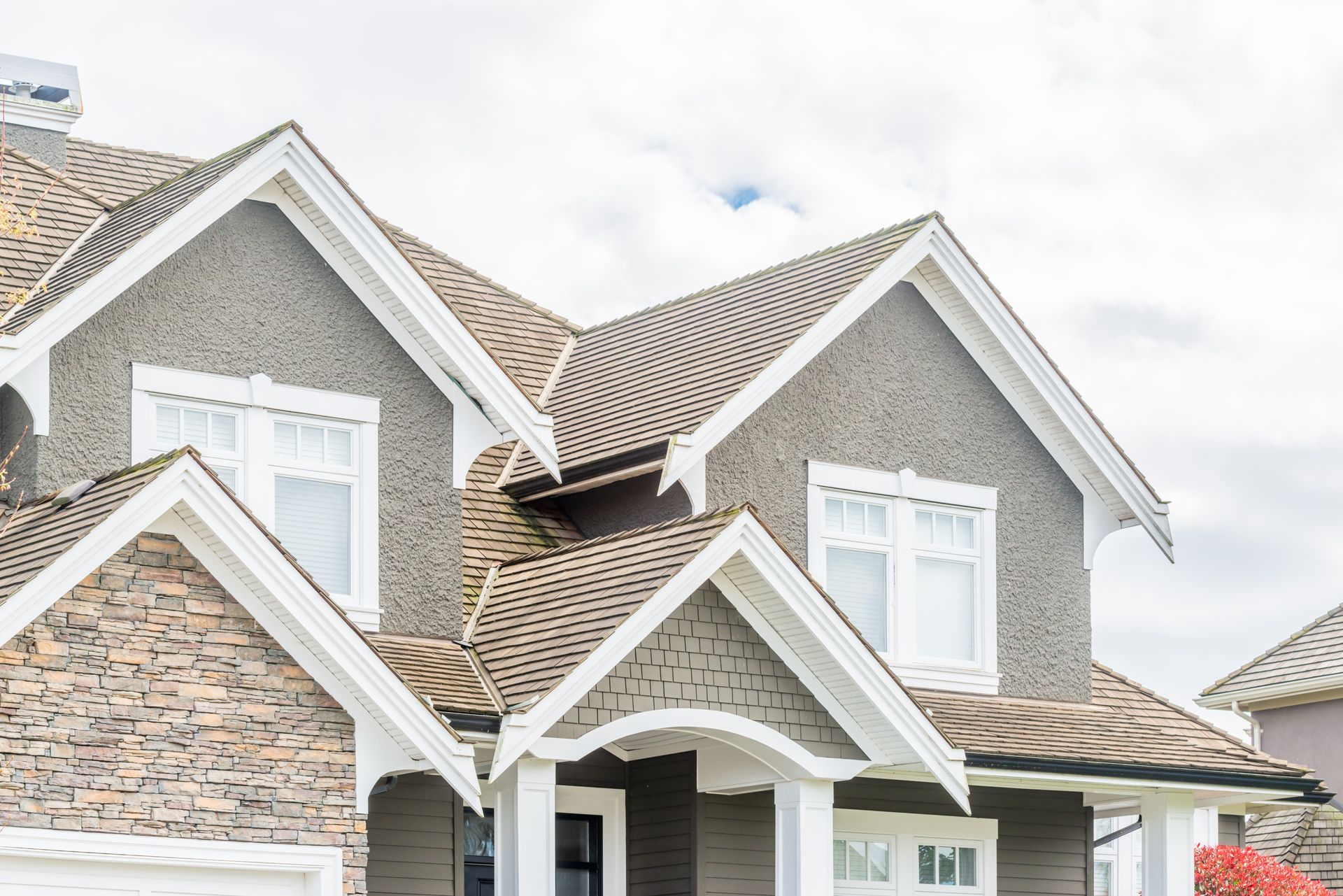 Multi-gabled house with gray siding, stone accents, white trim, and a brown shingle roof against a cloudy sky. Multi-gabled house with gray siding, stone accents, white trim, and a brown shingle roof against a cloudy sky.