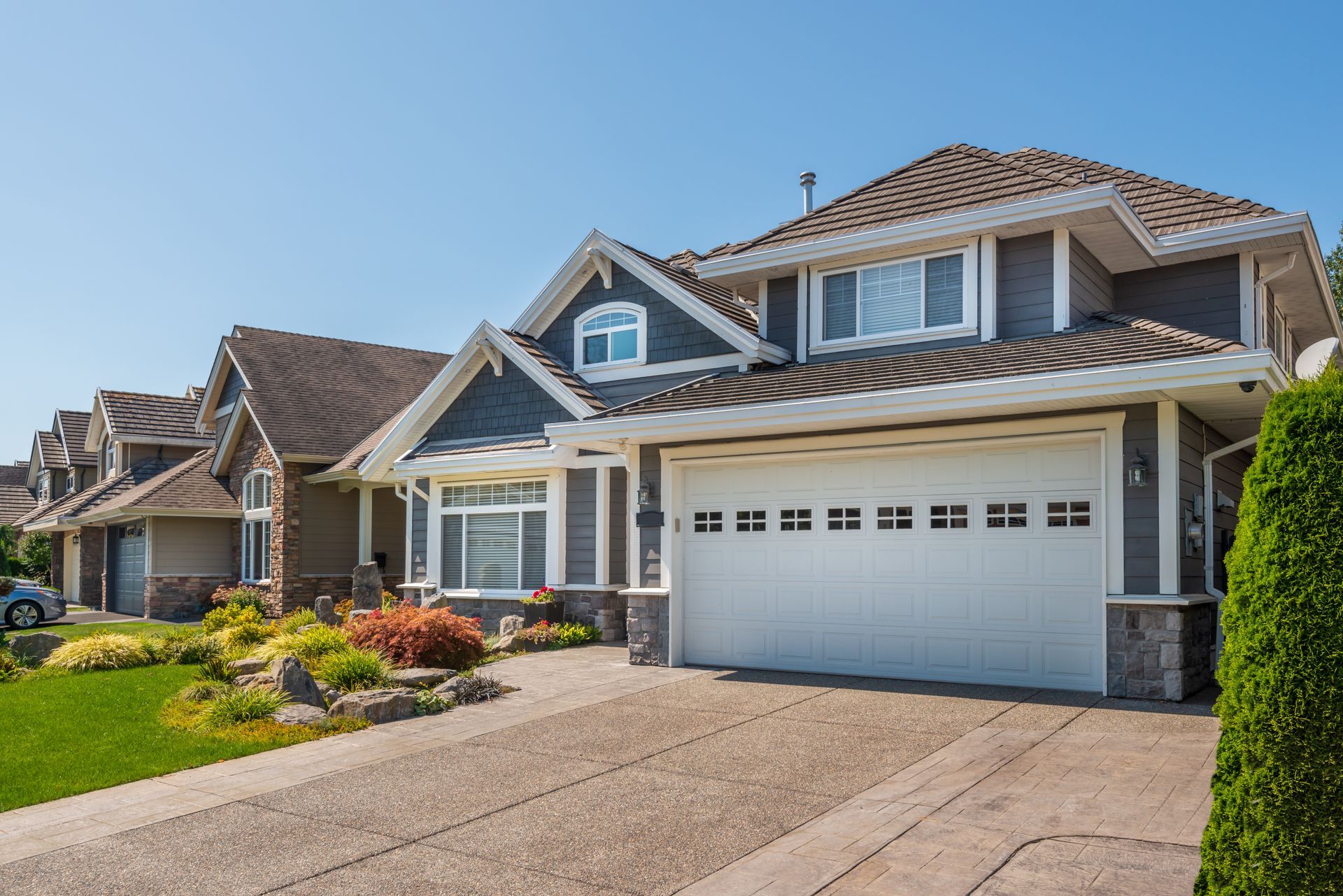 Two-story gray house with white garage door and concrete driveway. Sunny day with green grass and shrubs.