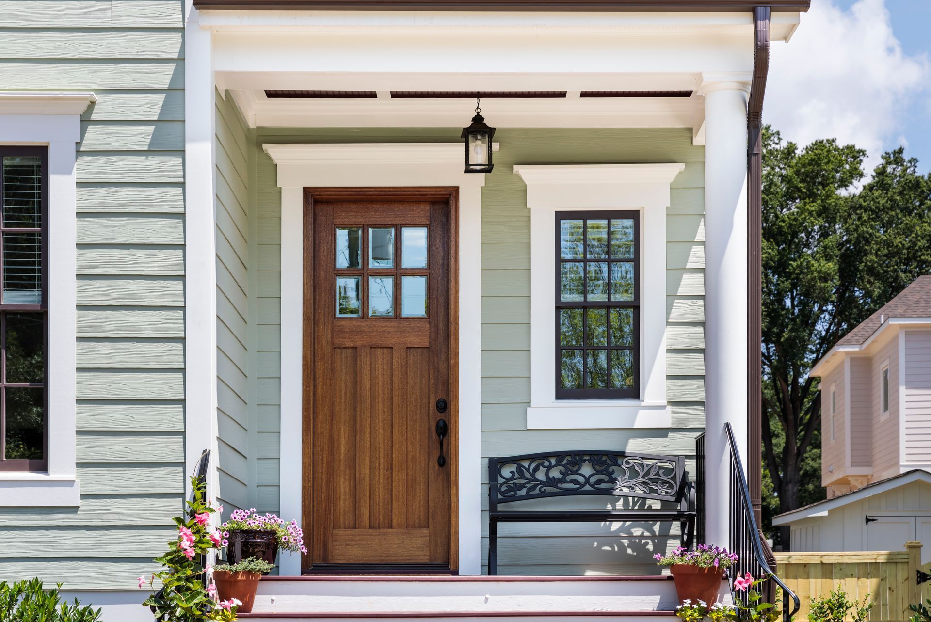 Wooden front door with glass panes, flanked by window and columns. Light green siding, porch bench, and potted flowers.