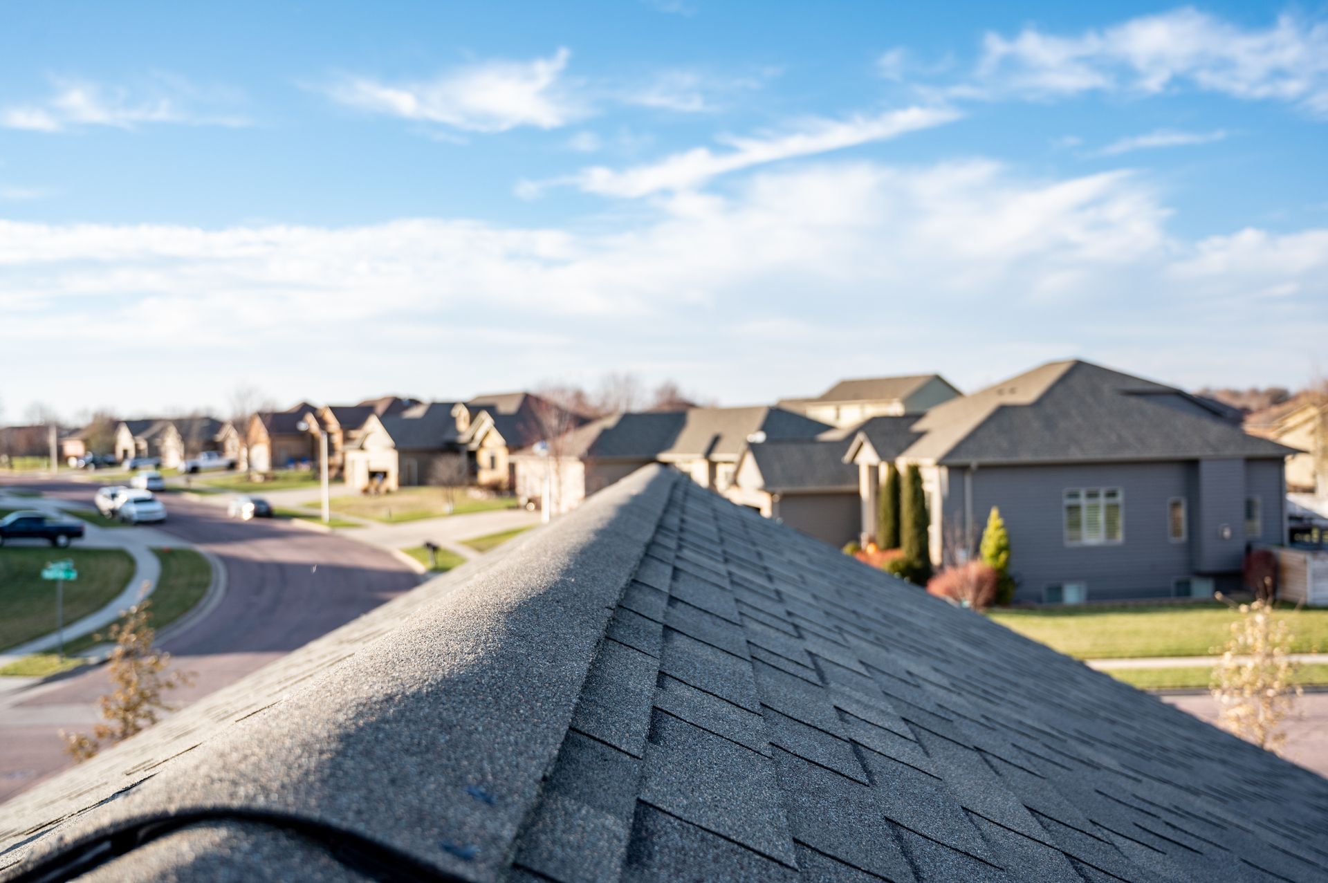 View from a roof of a neighborhood with houses, streets, and blue sky. The roof is textured asphalt shingles.
