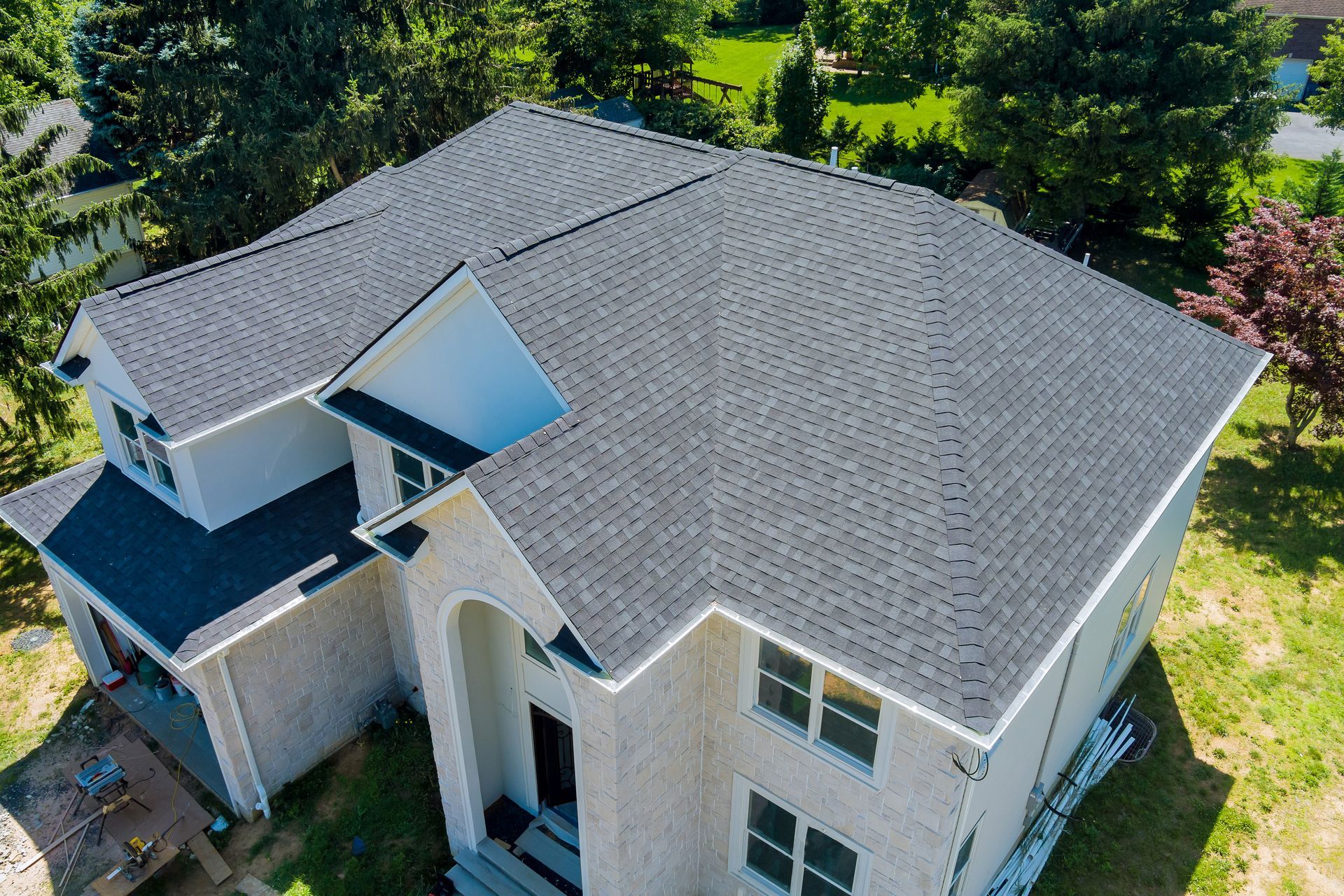 Gray asphalt shingle roof on a two-story beige house with surrounding green trees and grass.