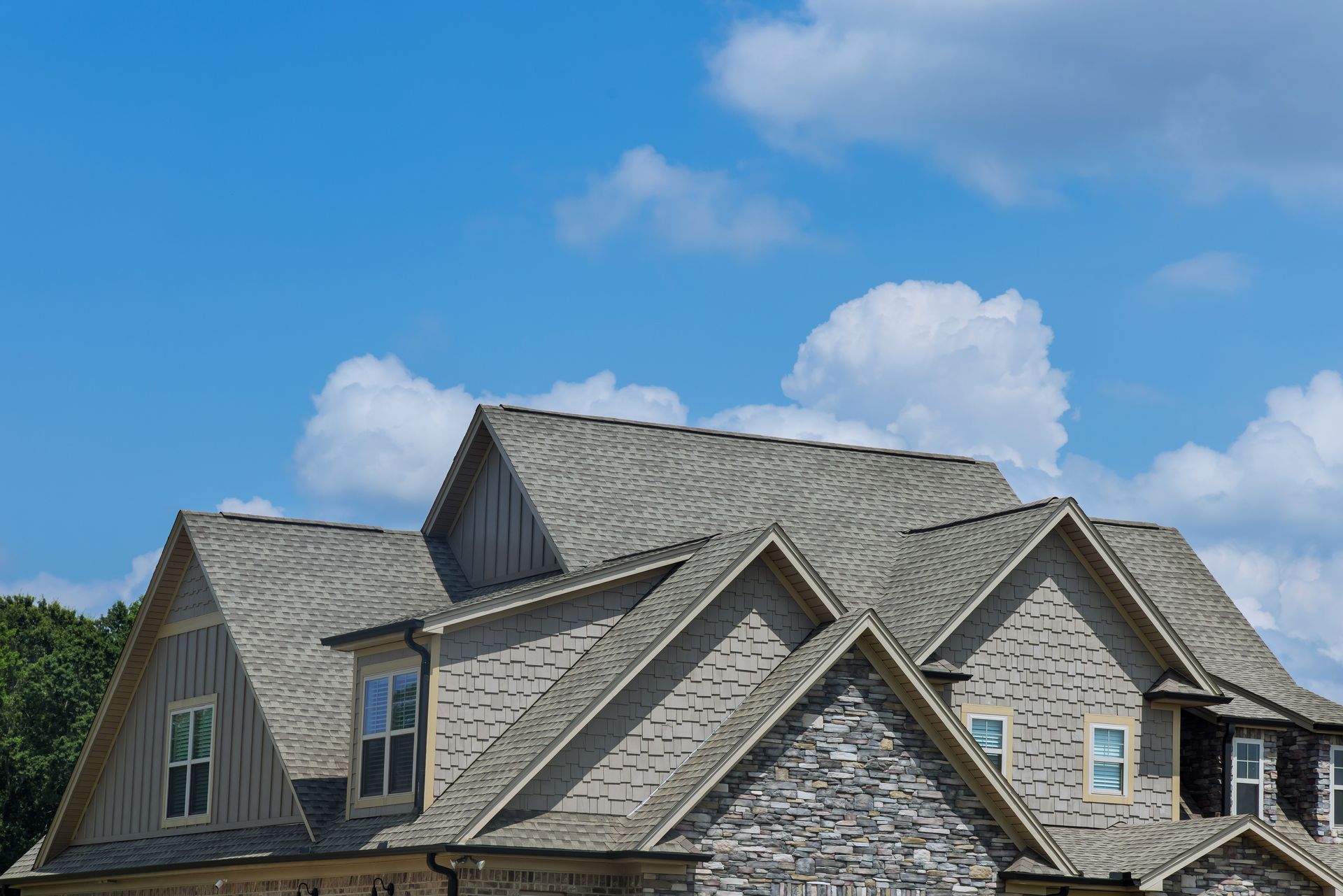 House with gray shingle roof, stone and wood siding, blue sky with clouds.