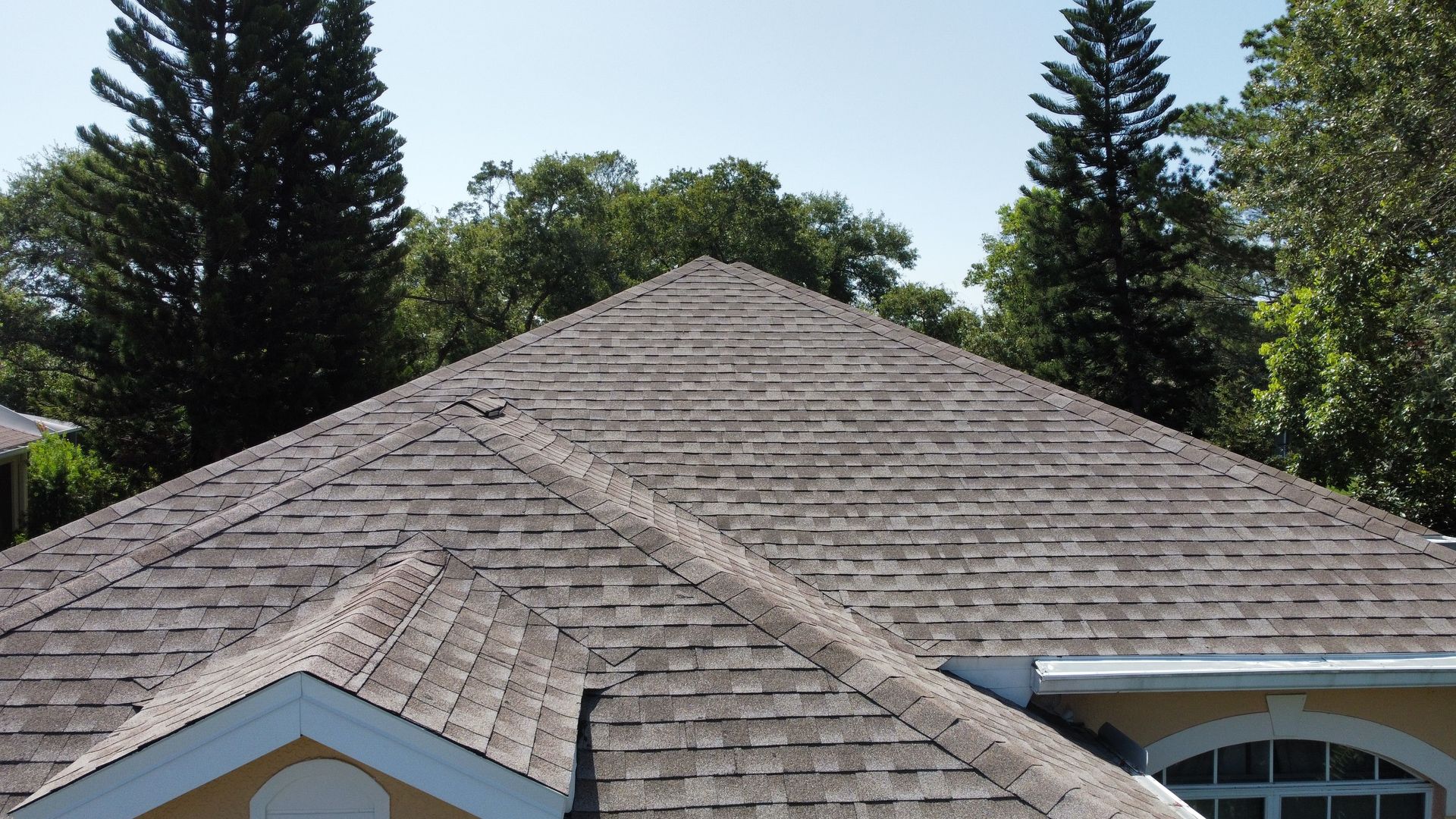 Gray shingled roof of a house, triangular shape, surrounded by green trees under a blue sky. Gray shingled roof of a house, triangular shape, surrounded by green trees under a blue sky.