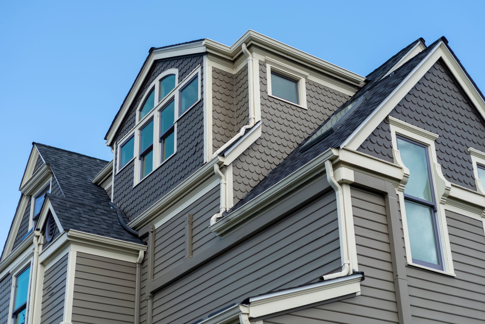 Grey and tan house with multiple dormers, windows, and dark roof against a clear blue sky.