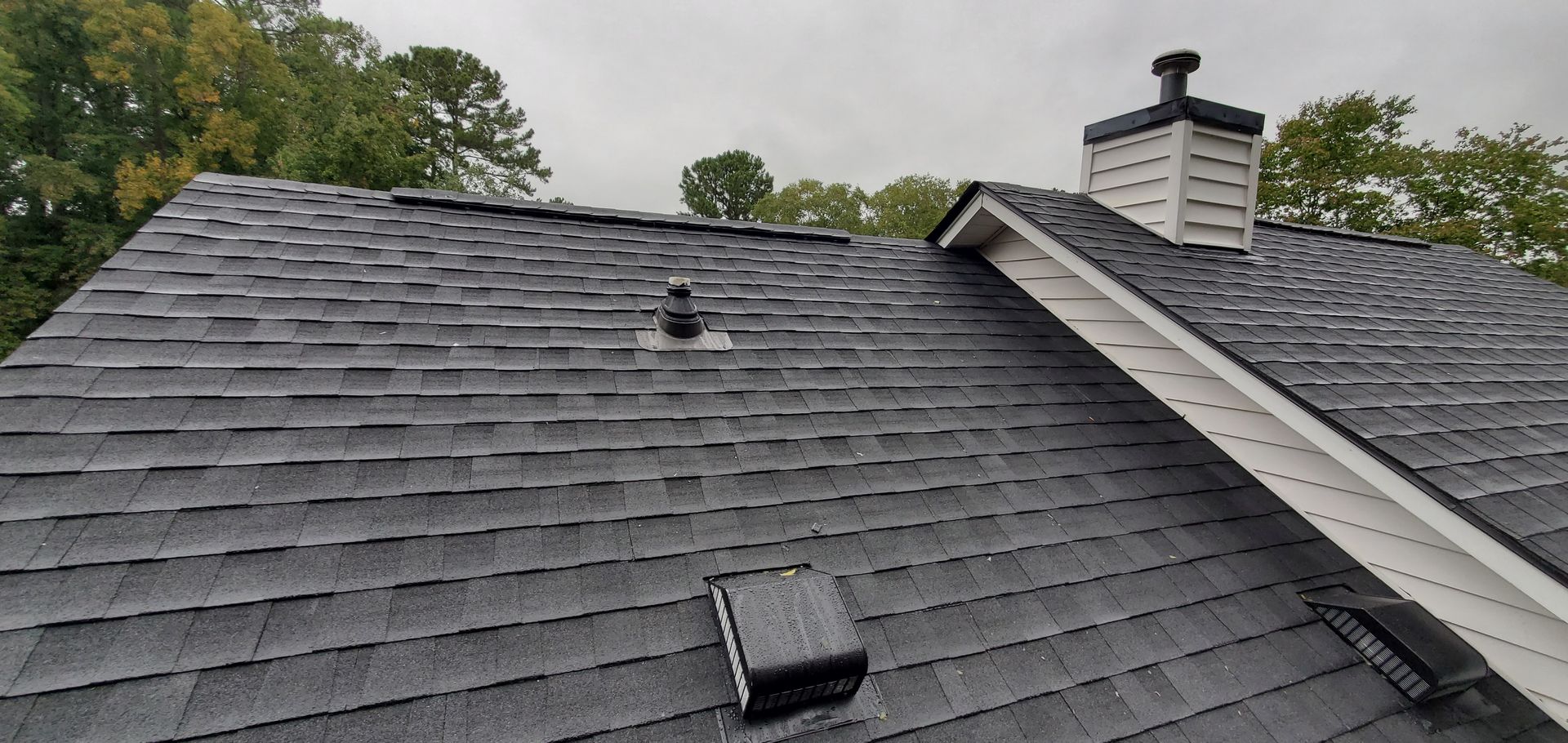 View of a dark gray shingle roof with a chimney and vent pipes, set against an overcast sky.