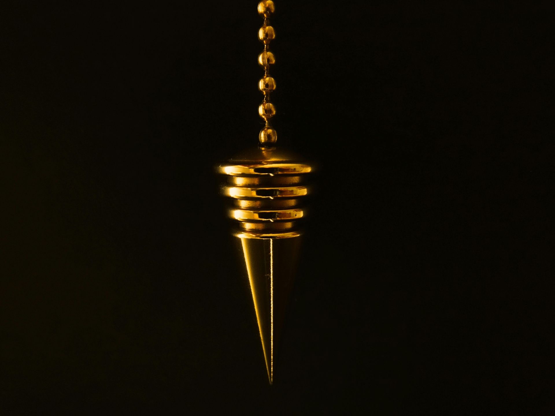 Golden pendulum with conical tip, suspended by a beaded chain against a black background.
