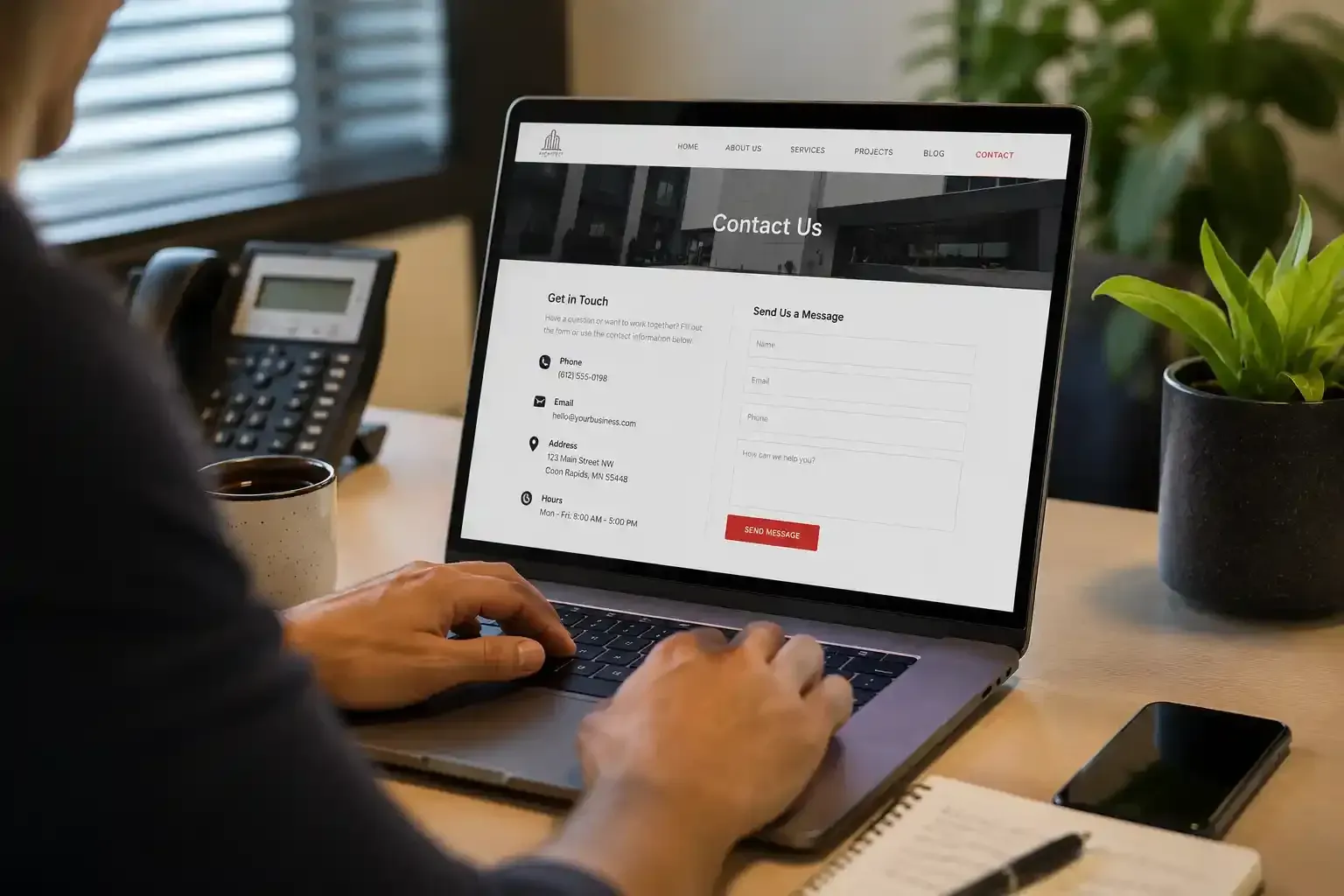 Person using a laptop looking at Contact Pag at a desk with a phone, plant, and office phone nearby.