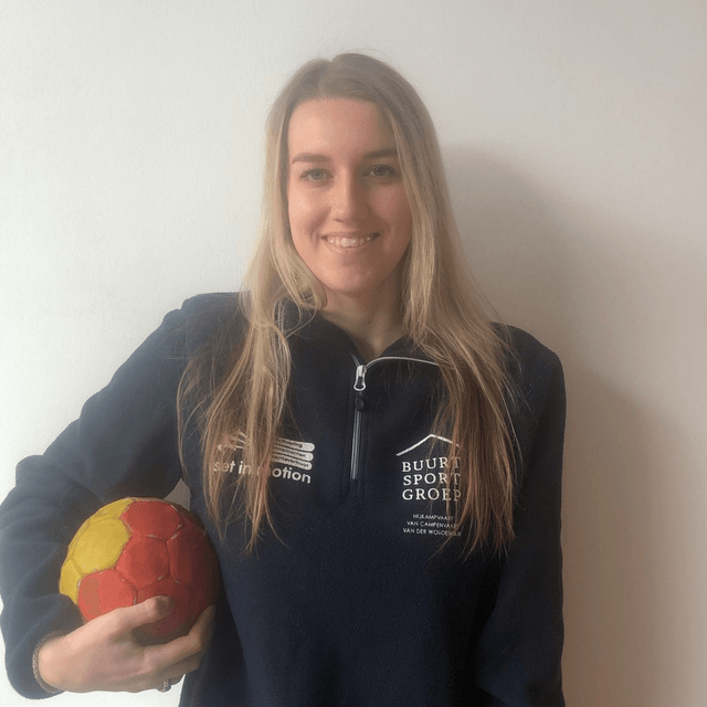 Vrouw met een handbal in haar handen, gekleed in een donkerblauwe trui, lachend. Tegen een witte muur.