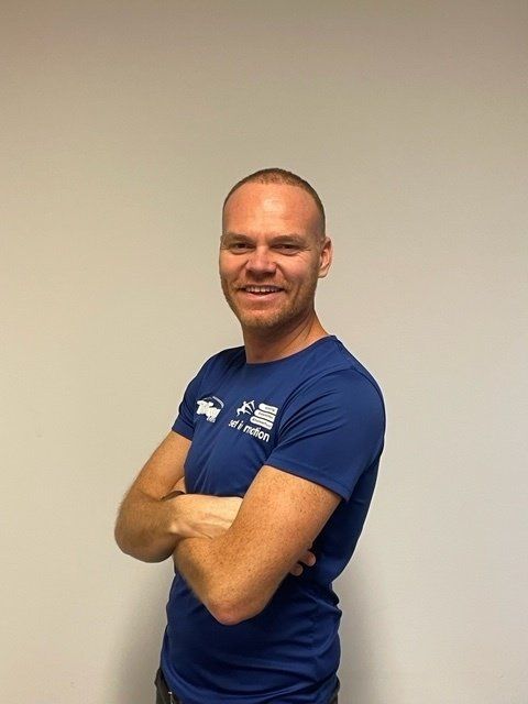 Een man met gekruiste armen, glimlachend, gekleed in een blauw shirt met logo, staat voor een witte muur.