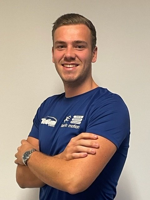 Een glimlachende man met gekruiste armen, gekleed in een blauw T-shirt en met een horloge om.