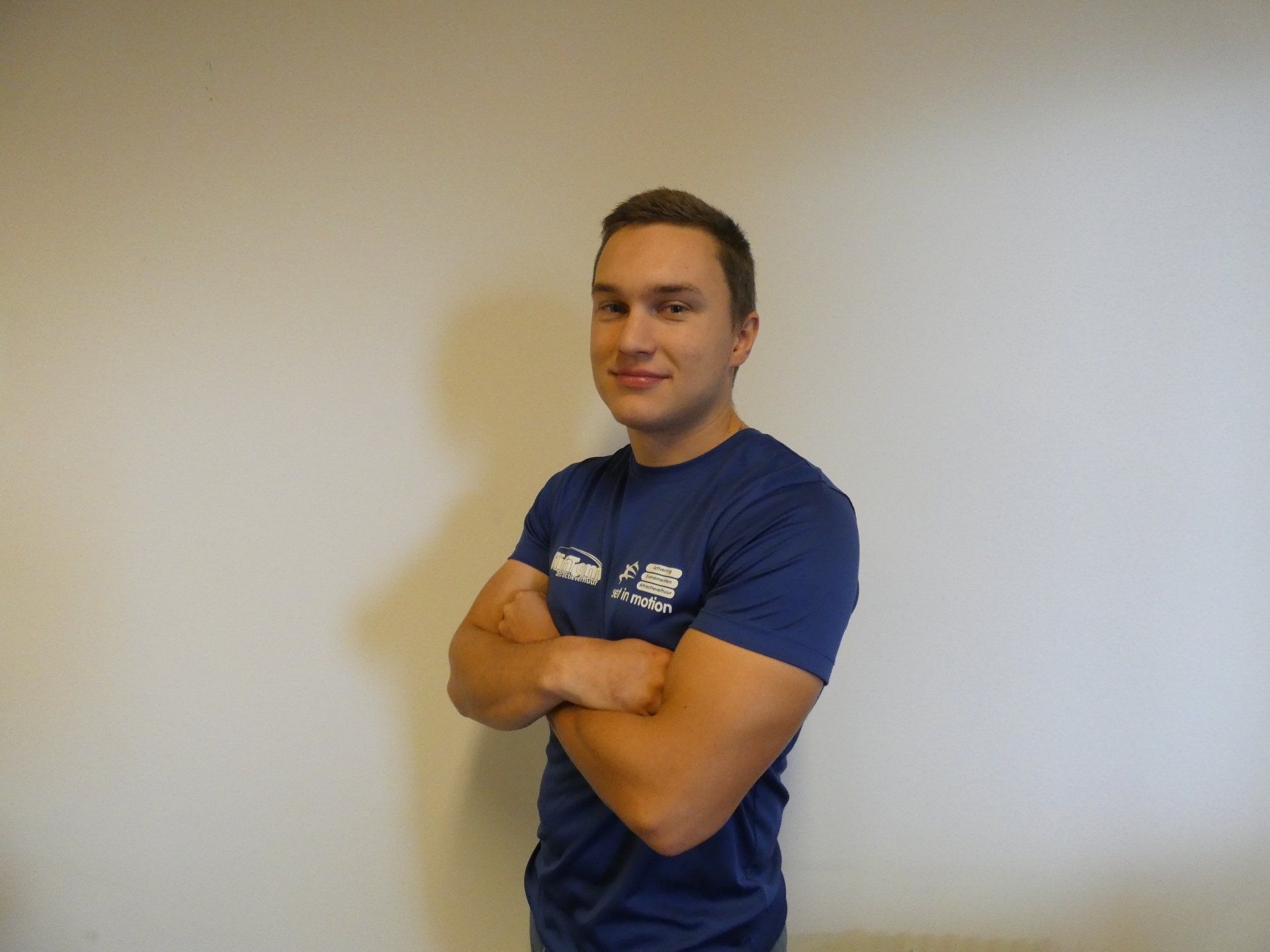 Een man in een blauw shirt, met de armen over elkaar, glimlacht. Hij staat tegen een effen muur.