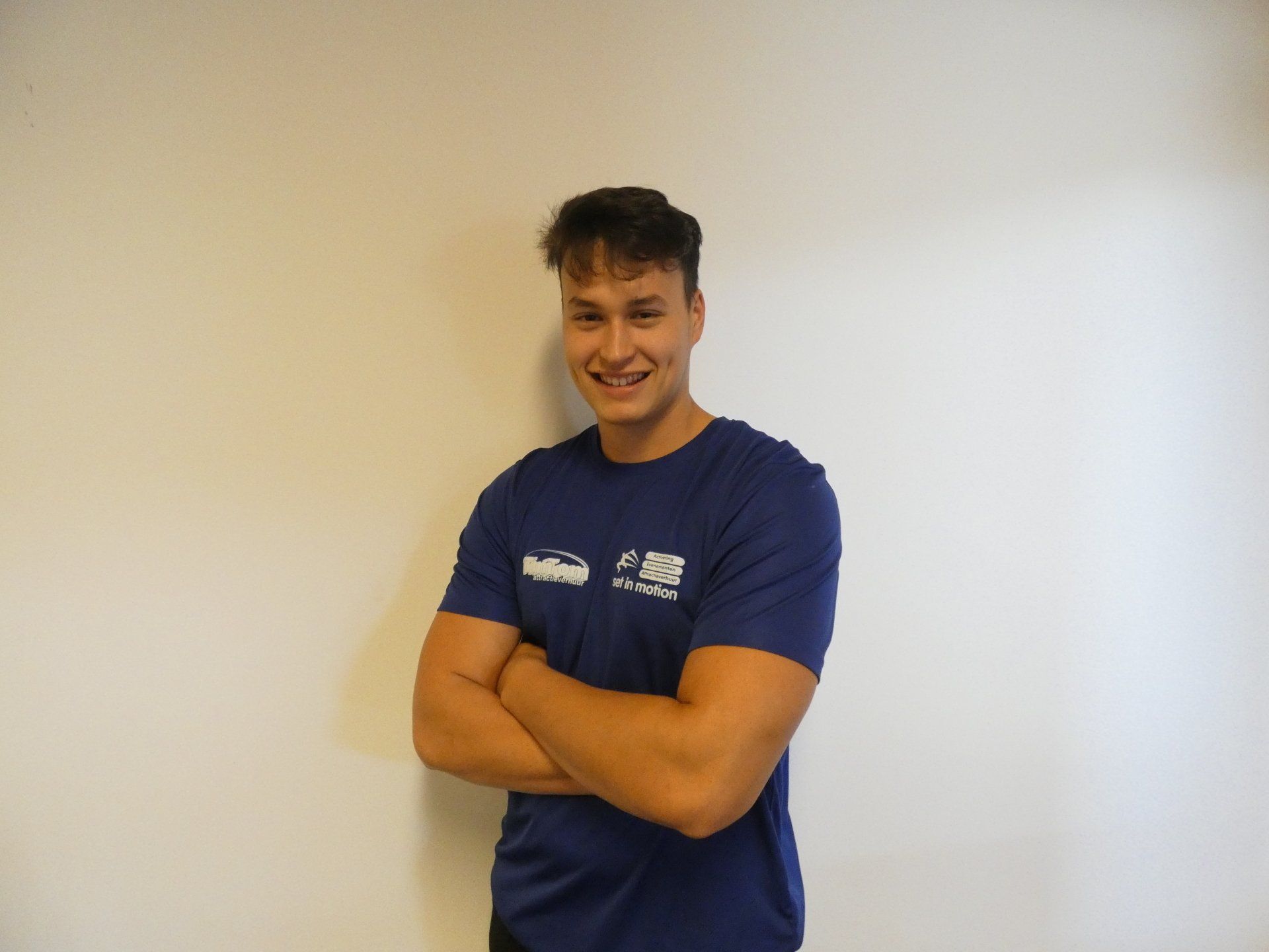 Een man in een blauw T-shirt met gekruiste armen lacht tegen een witte muur.