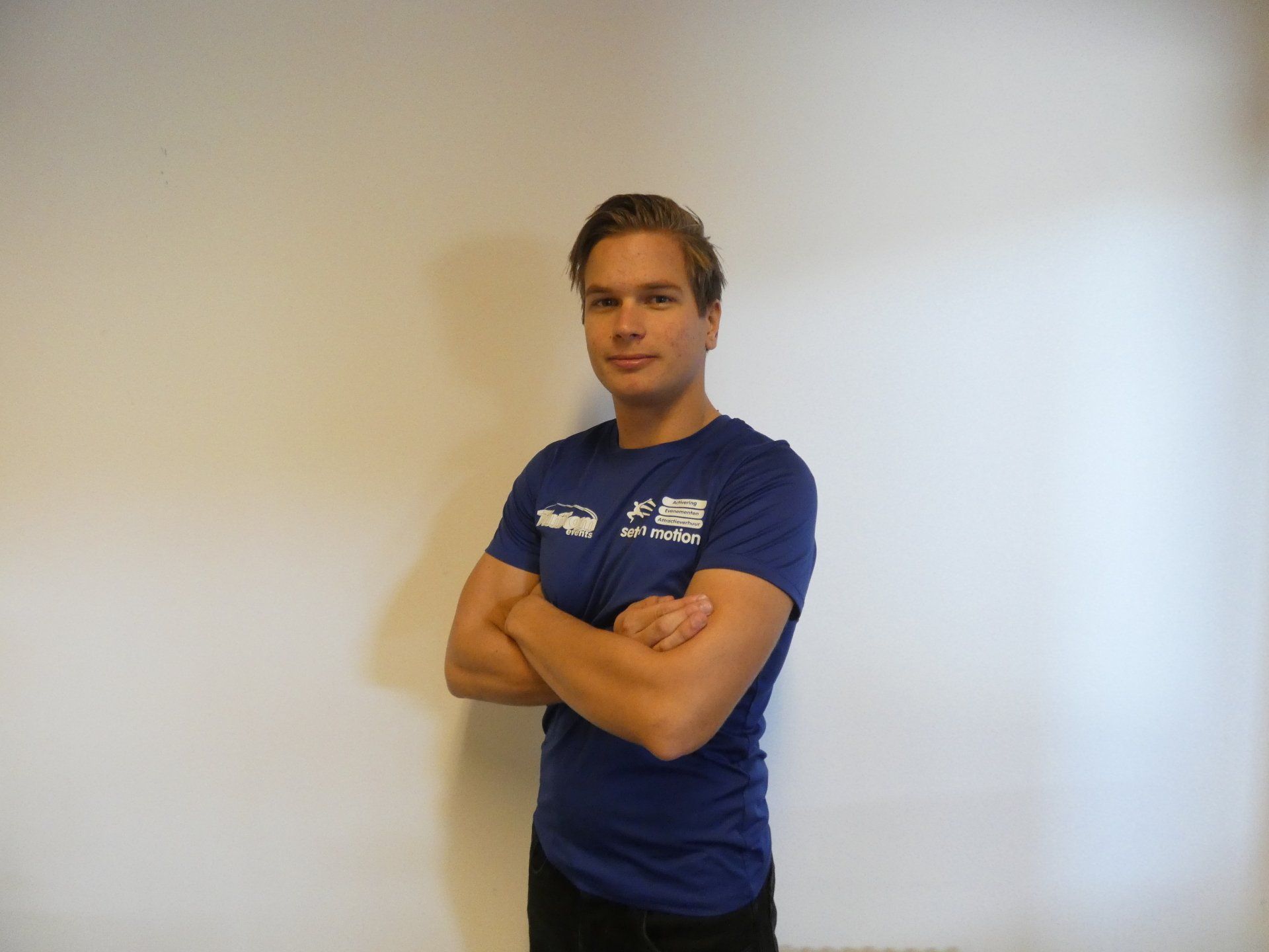 Een man met gekruiste armen, gekleed in een blauw shirt en een zwarte broek, staat voor een witte muur.