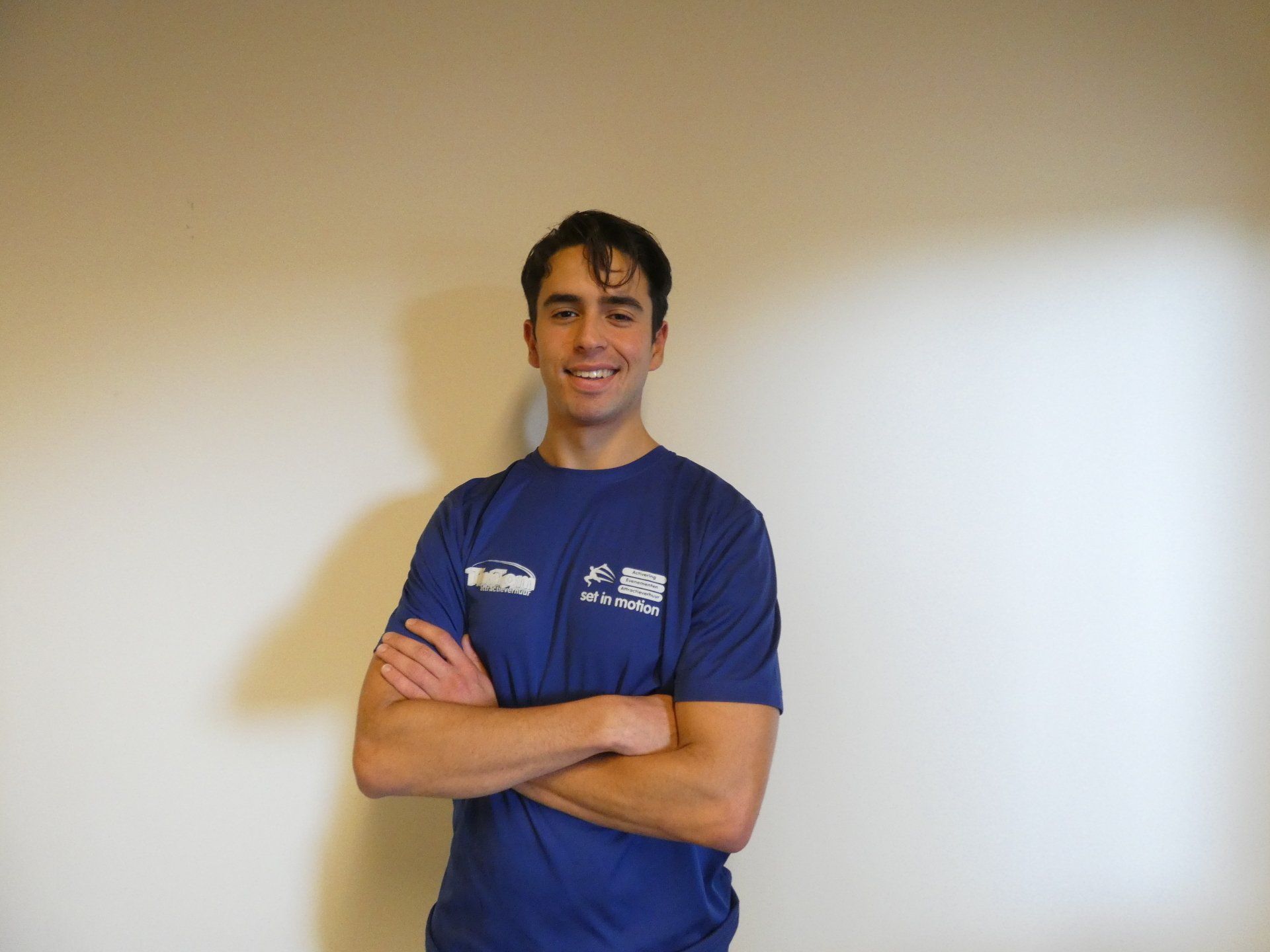 Een man met donker haar en een blauw shirt staat met gekruiste armen, glimlachend, tegen een beige muur.