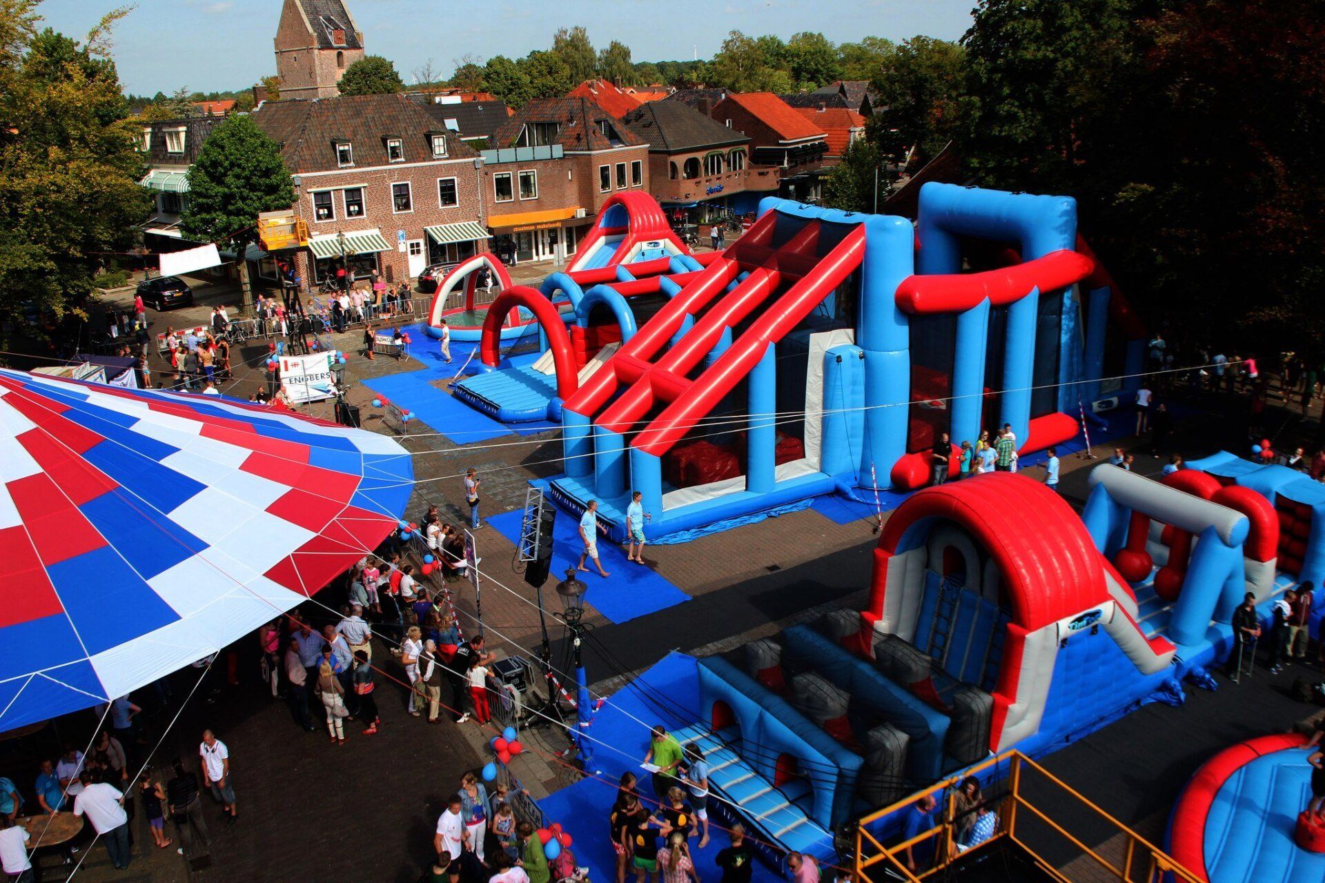 Groot opblaasbaar hindernisparcours op een buitenevenement met veel publiek. Rood, wit en blauw van kleur. Er is een grote menigte bijeen.