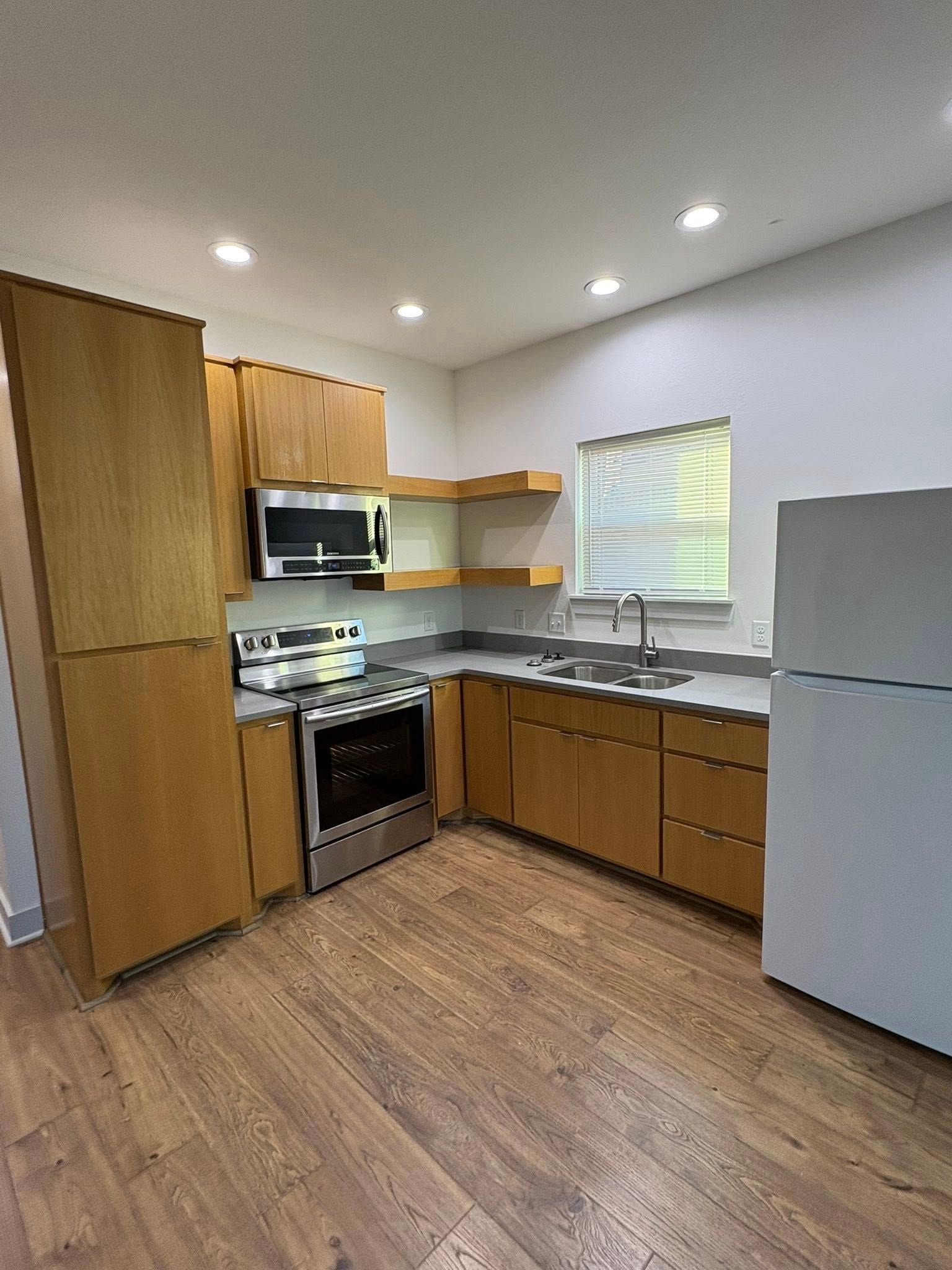 Small kitchen with wood cabinets, gray fridge, stove, sink, and wooden floor under recessed lights.