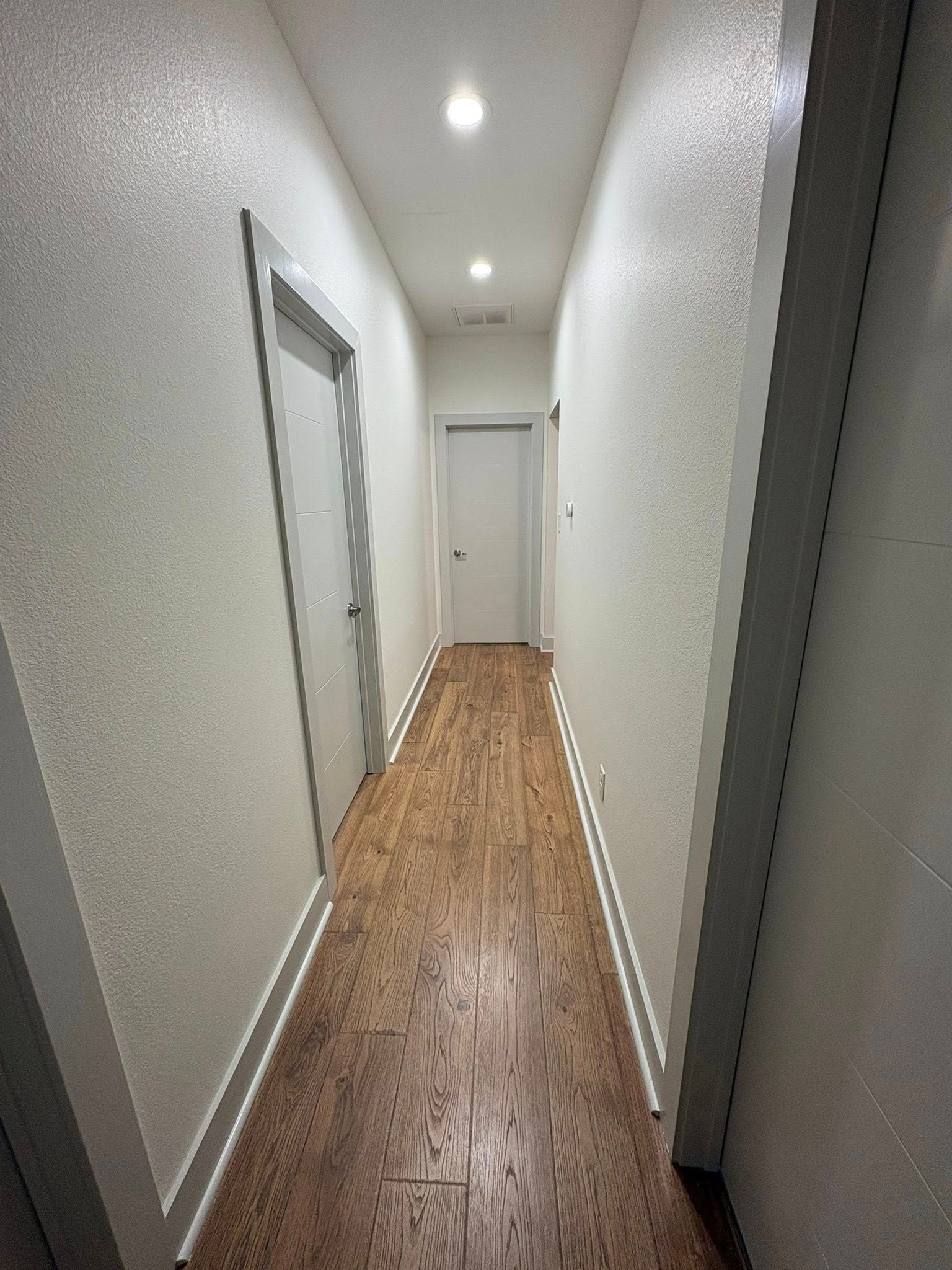 Narrow hallway with white walls, wood floor, and ceiling lights, leading to a closed door.