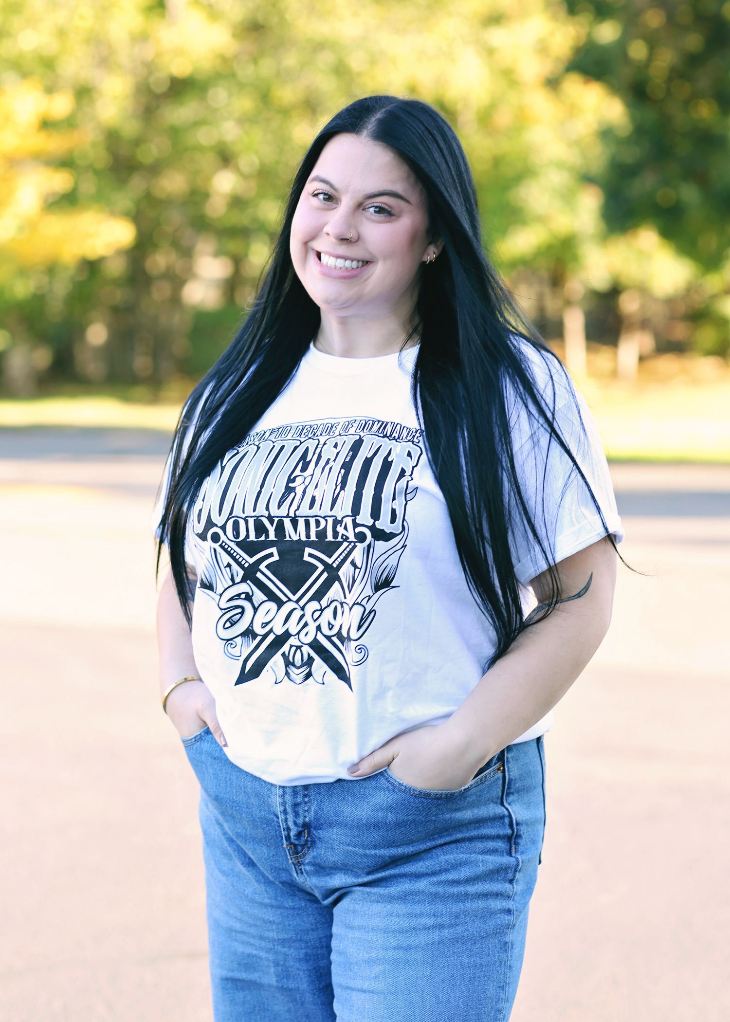 Woman with long black hair, smiling, wearing a white graphic tee and jeans, standing outside with hands in pockets.