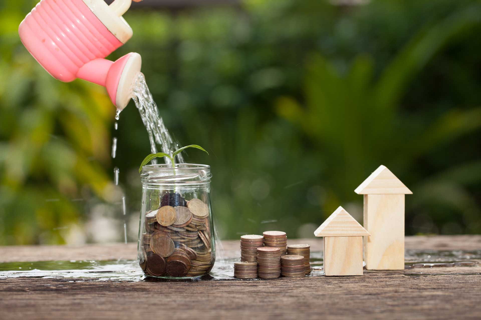 Pink watering can waters coins in a jar, with stacked coins and wooden house cutouts on a table, outdoors.